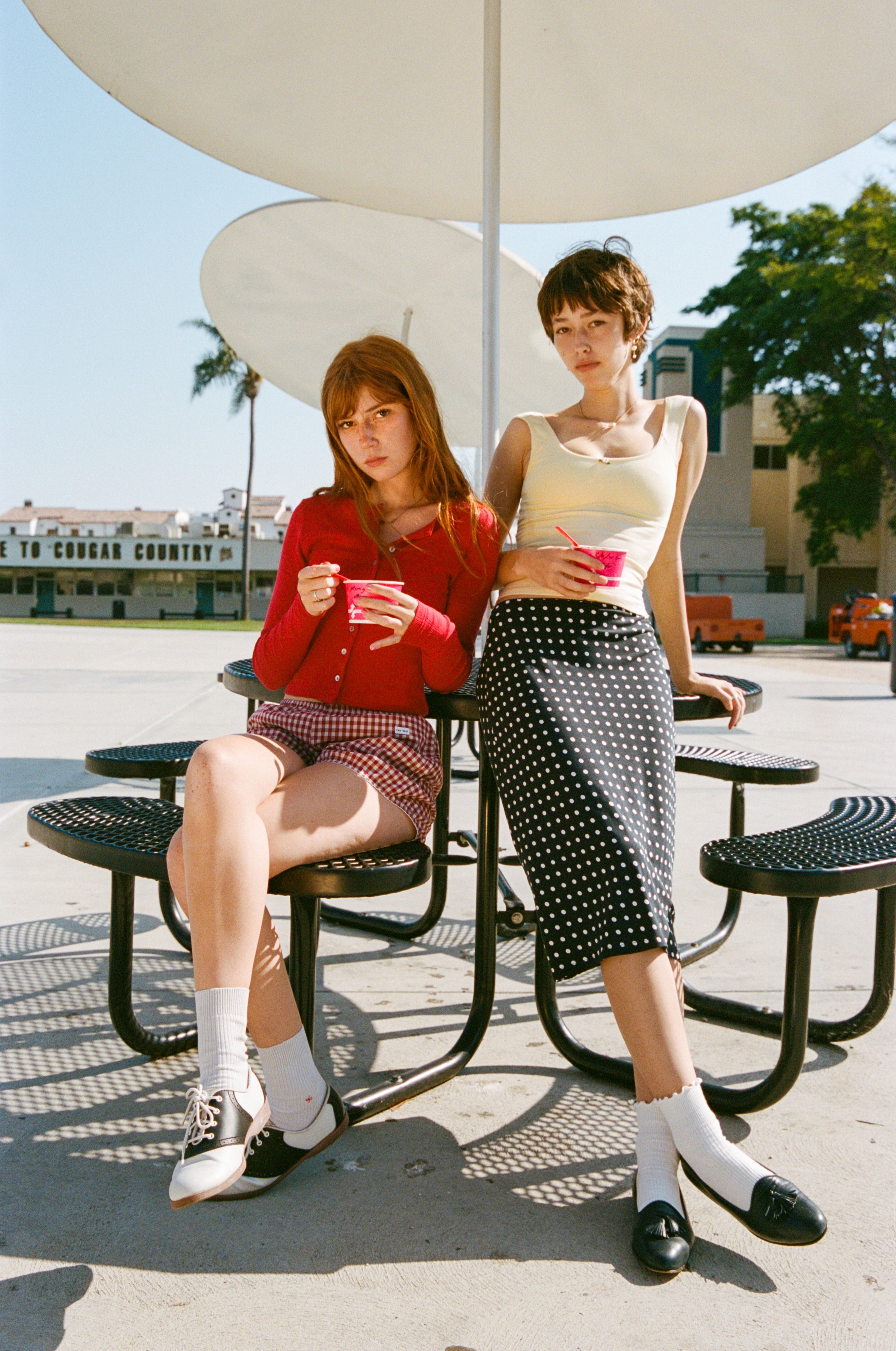 Two women sitting at outdoor table under umbrella wearing vintage-inspired outfits with polka dot patterns and retro styling