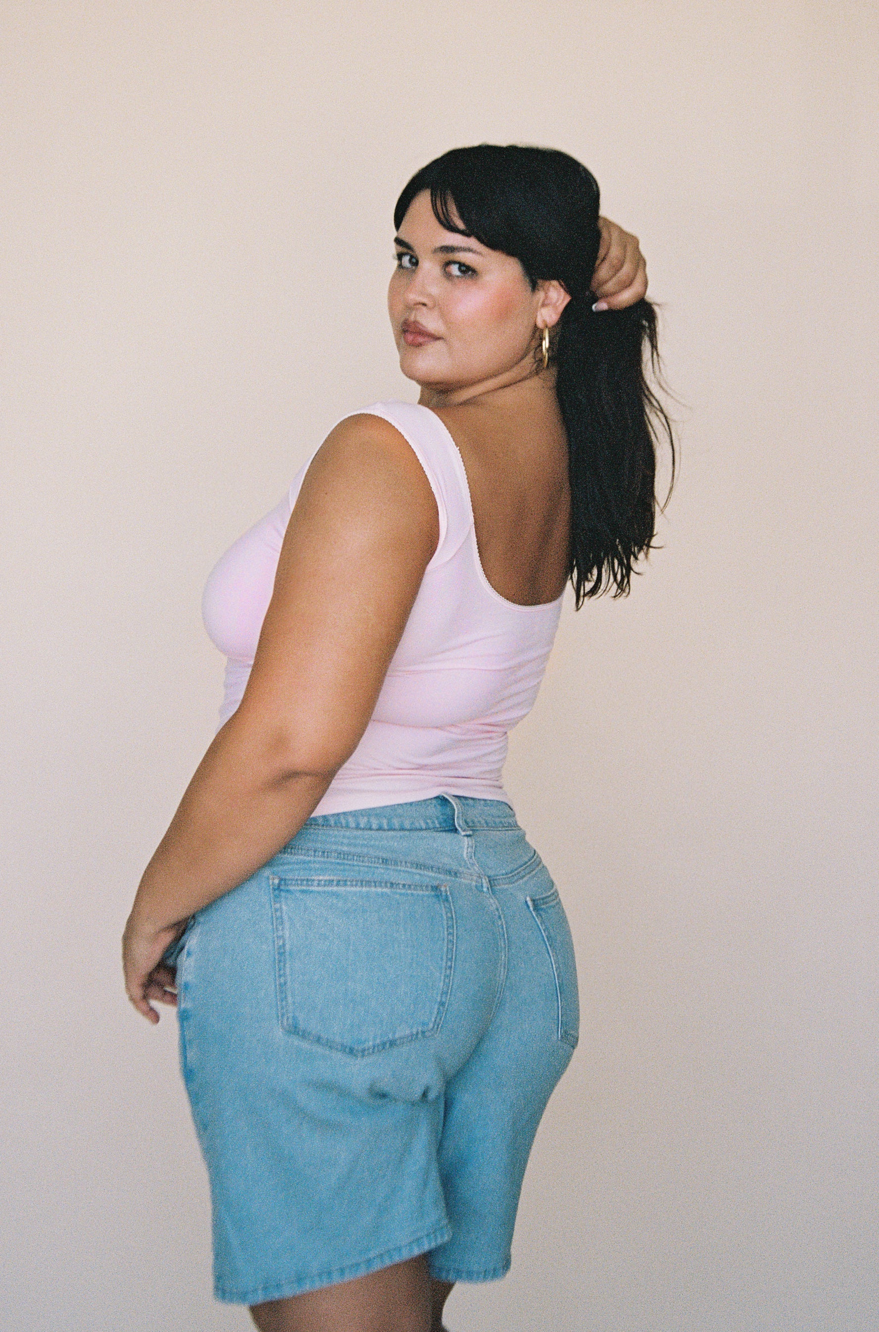 Woman wearing light blue high-waisted denim shorts with back pockets, paired with a white tank top against beige background