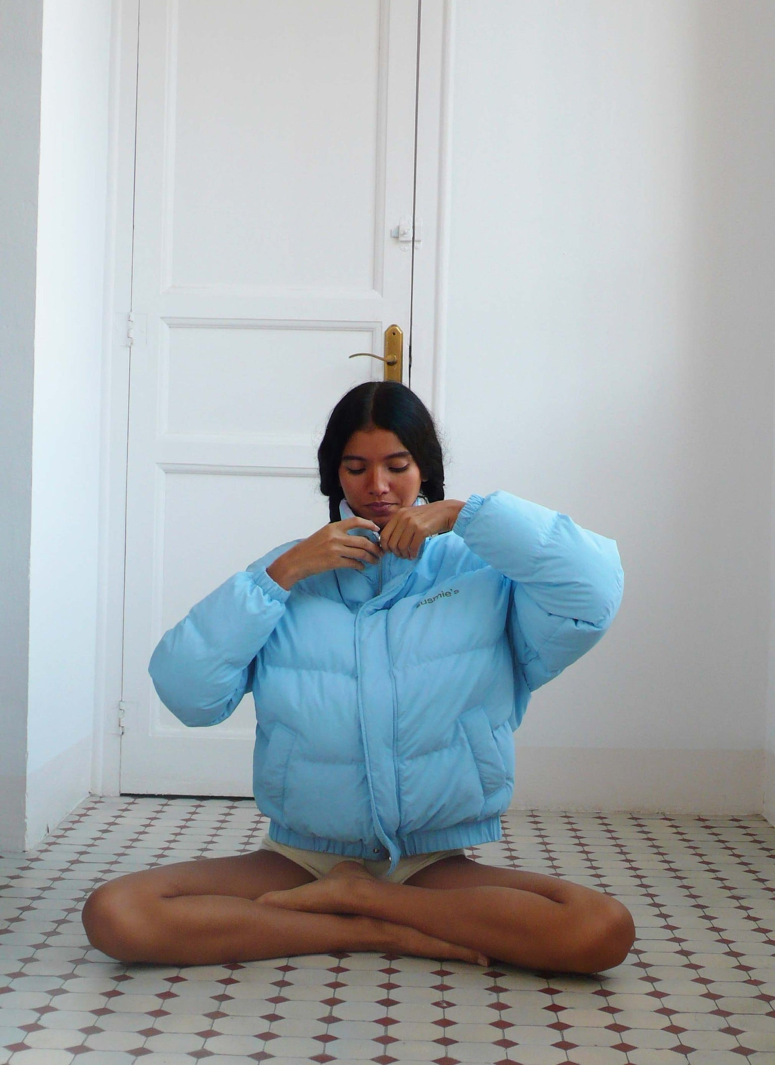 Woman wearing light blue puffer jacket while sitting cross-legged on patterned tile floor against white door