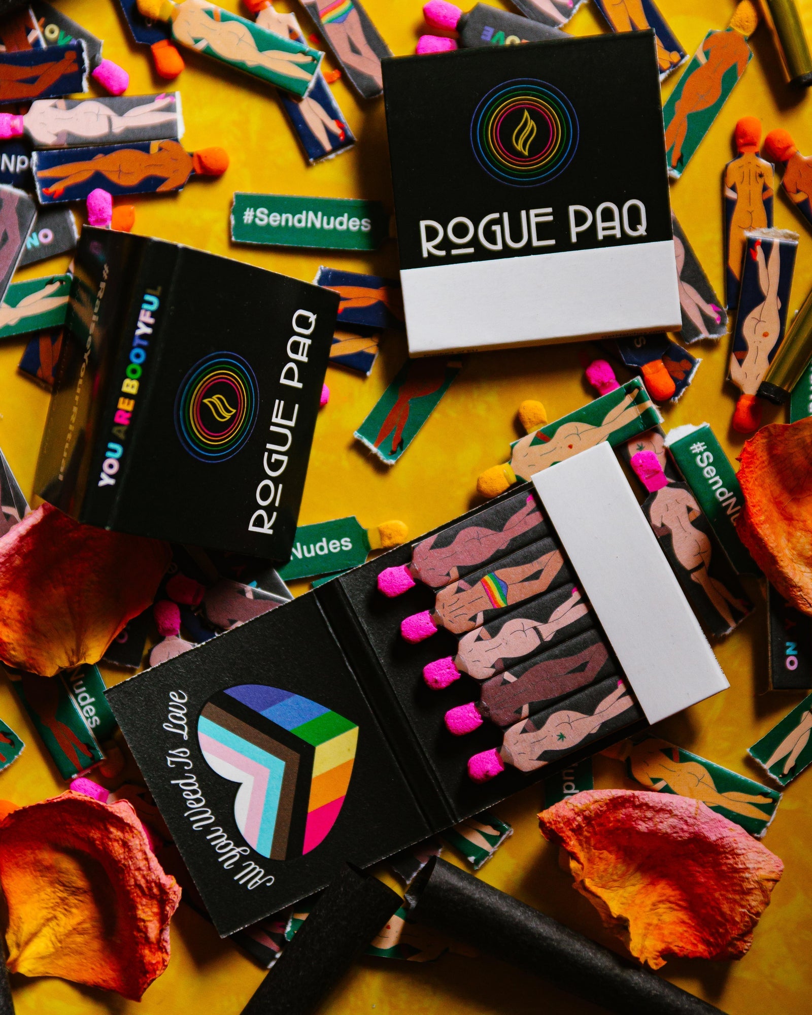 Colorful matchbook-style product packages for Rogue Paq scattered on yellow surface with illustrated figures and #SendNudes hashtag