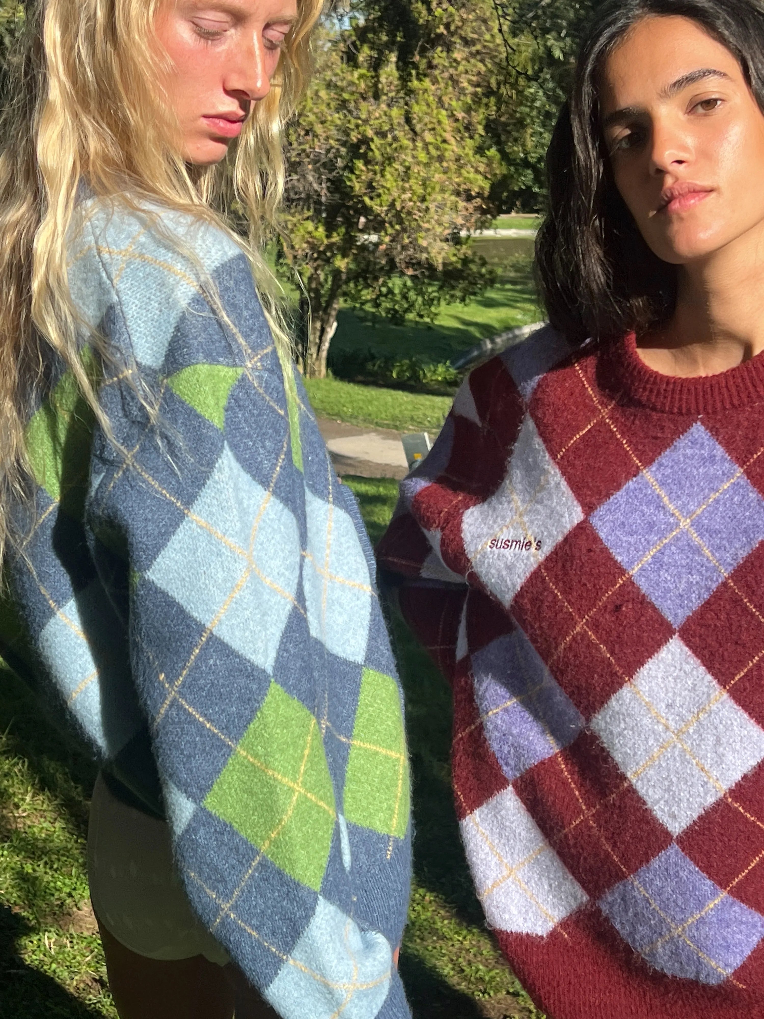 Two women wearing colorful argyle knit sweaters in blue-green and burgundy-purple tones, standing outdoors in natural light