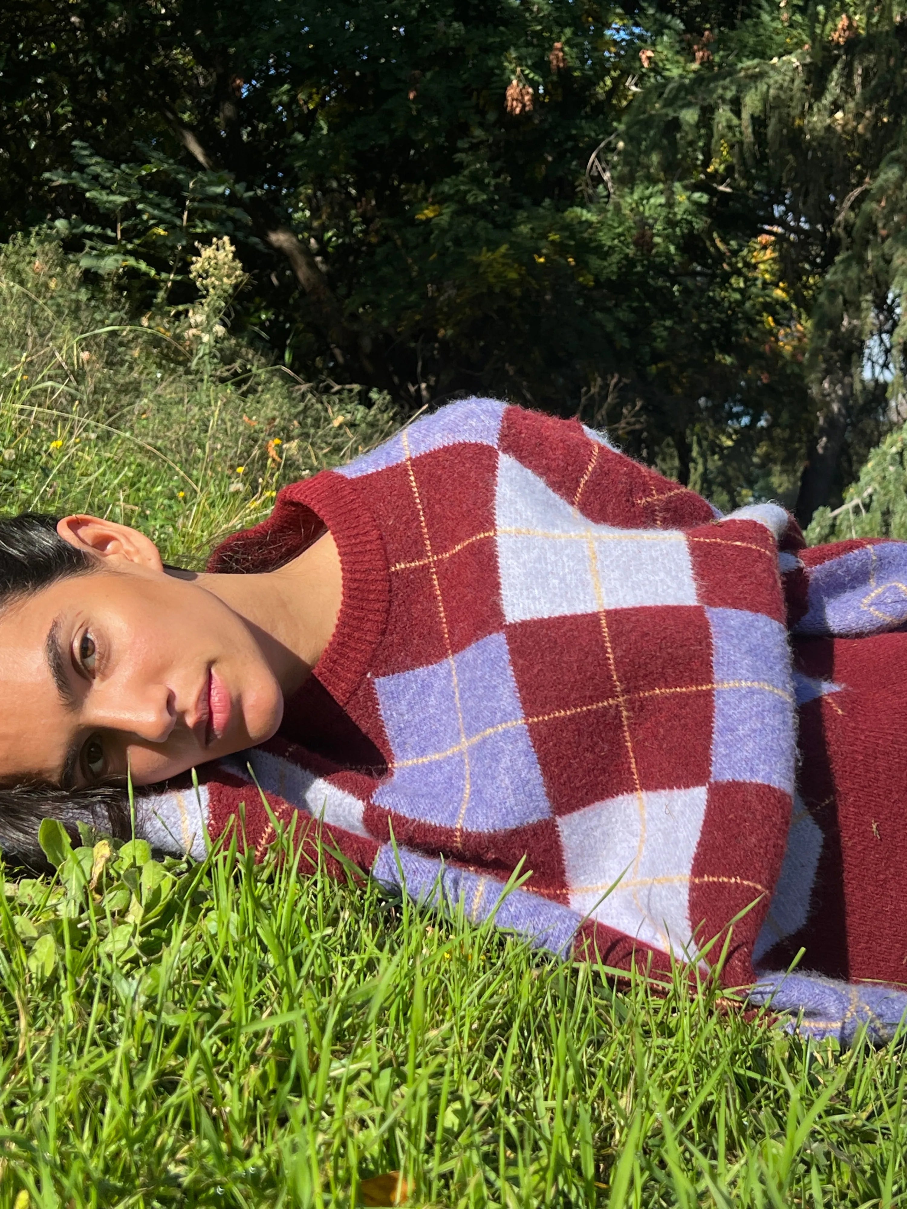 Woman wearing burgundy, lavender and white checkered knit sweater lying on grass in sunlight