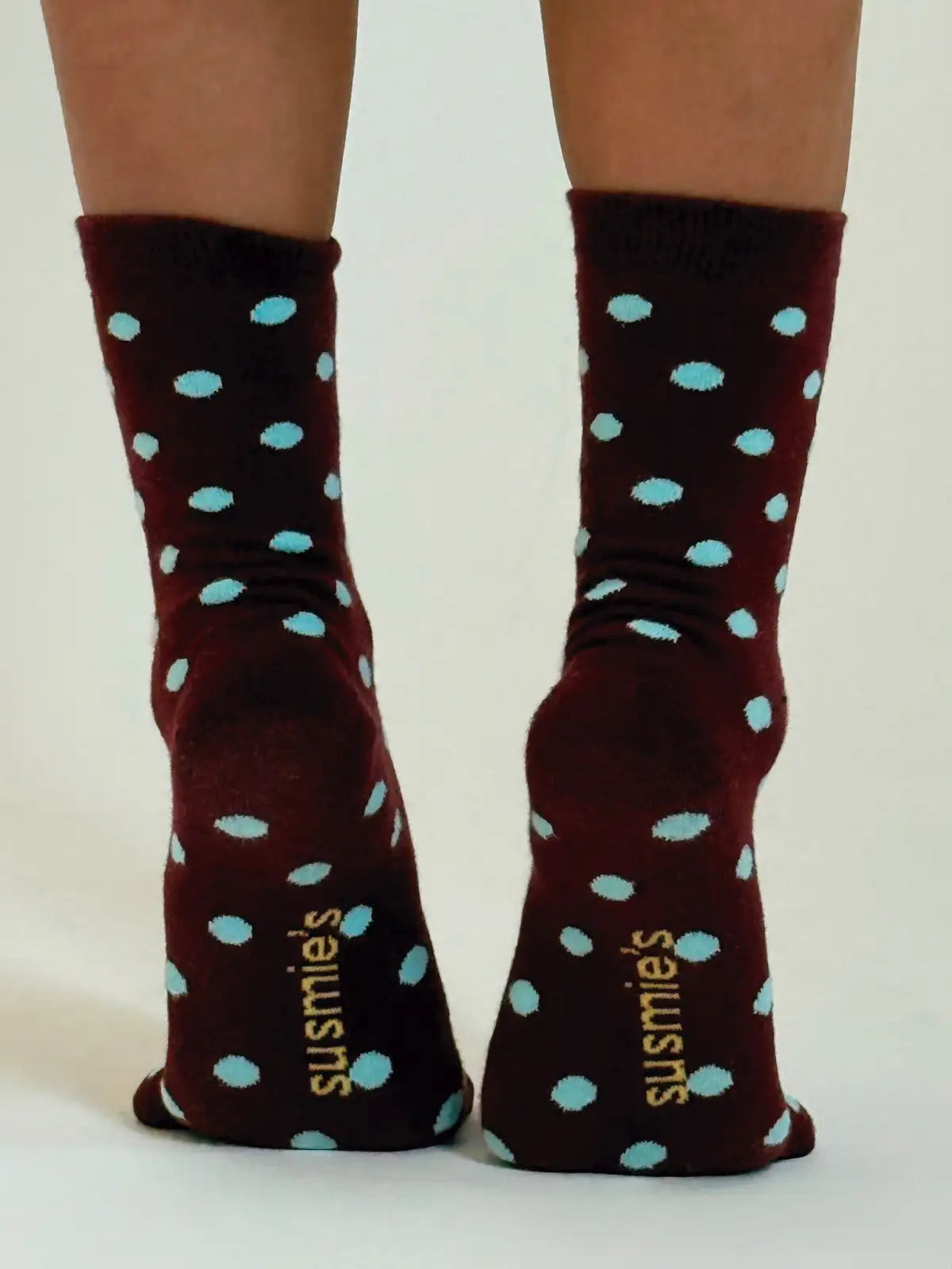 Dark burgundy socks with light blue polka dots and gold 'susmies' text on soles, shown on legs from behind