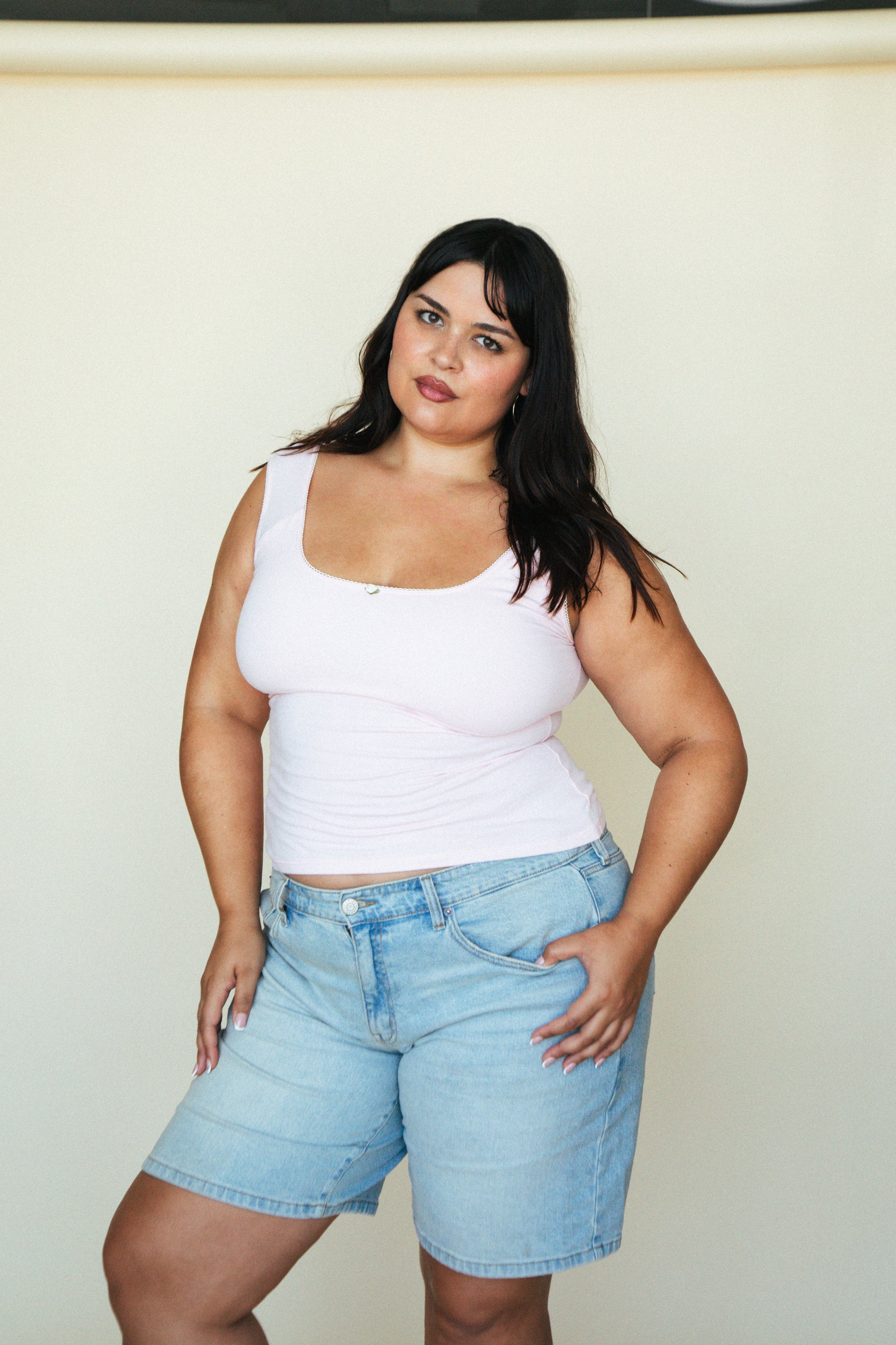 Plus-size woman wearing light blue denim Bermuda shorts and white tank top against cream background