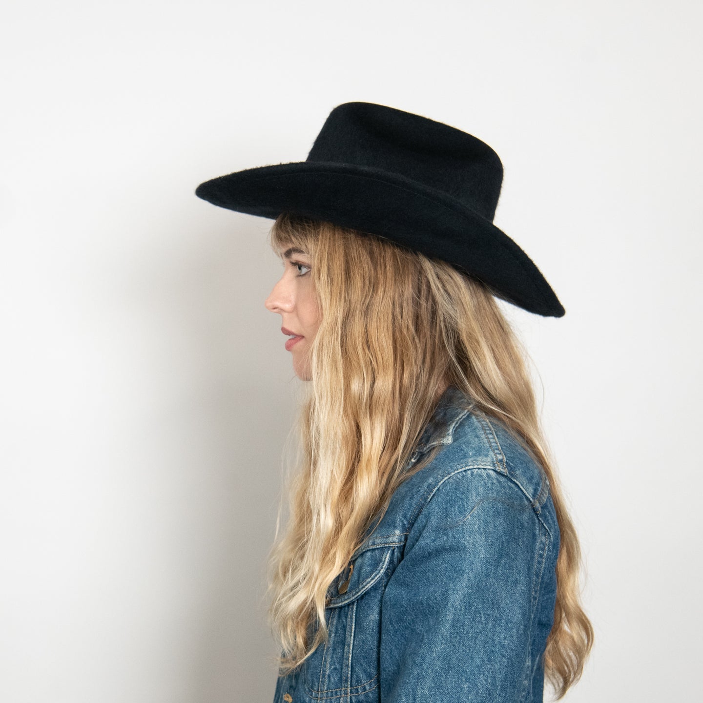 Black wide-brim felt fedora hat worn by blonde woman in denim jacket, shown in profile against white background