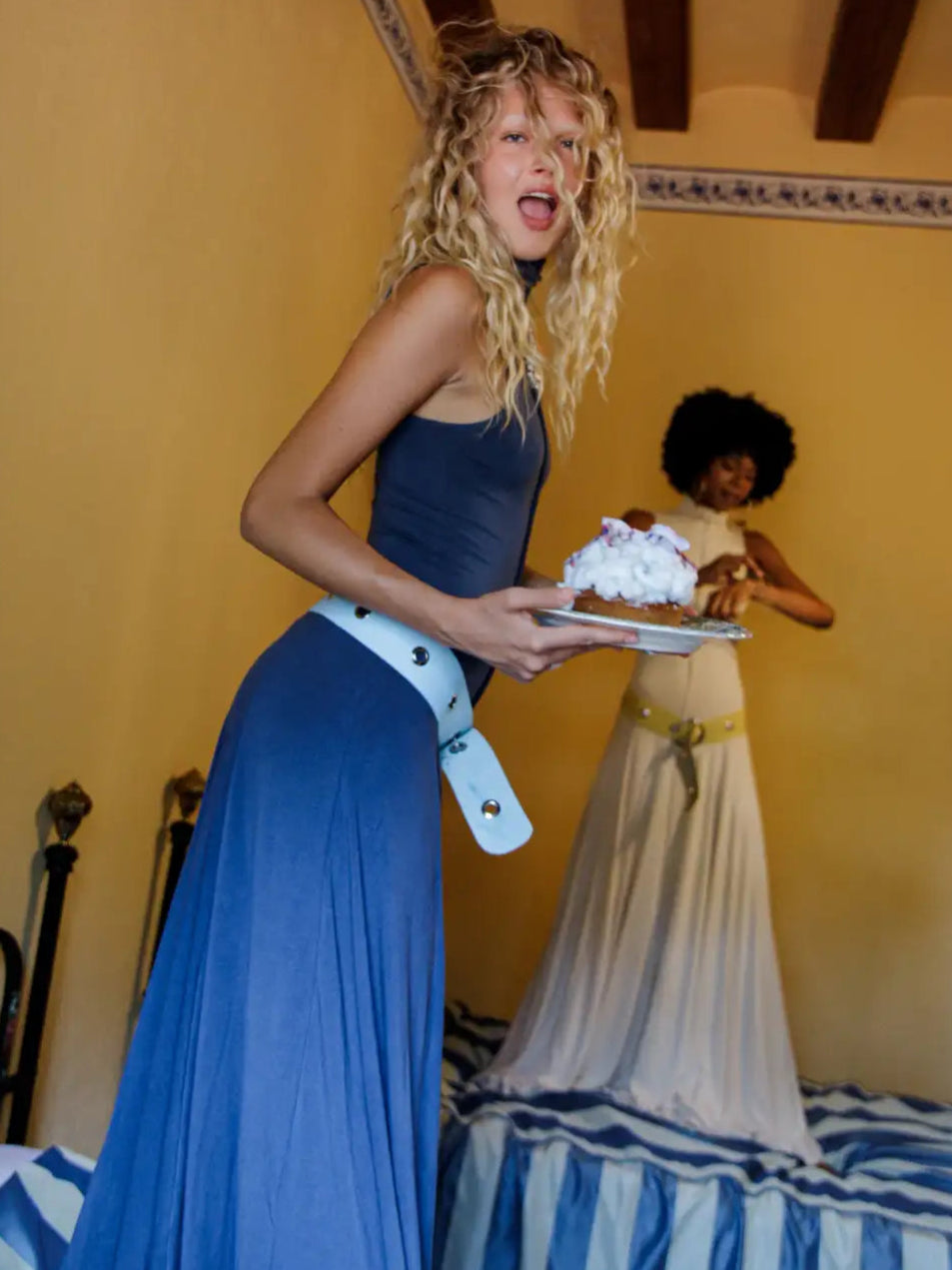 Woman in blue sleeveless pleated maxi dress with white belt detail holding a plate, featuring a halter neckline and flowing skirt.