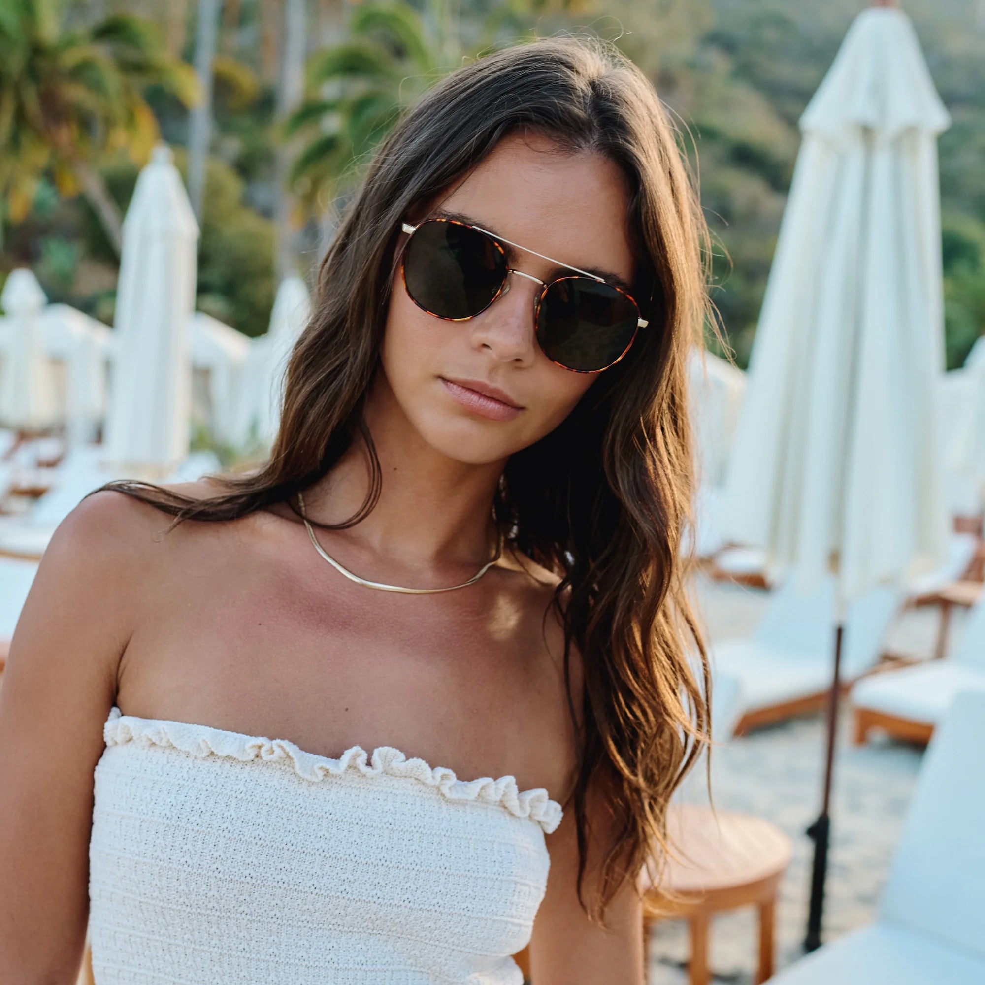 Woman wearing sunglasses and a white strapless top in a tropical setting with white umbrellas and greenery.