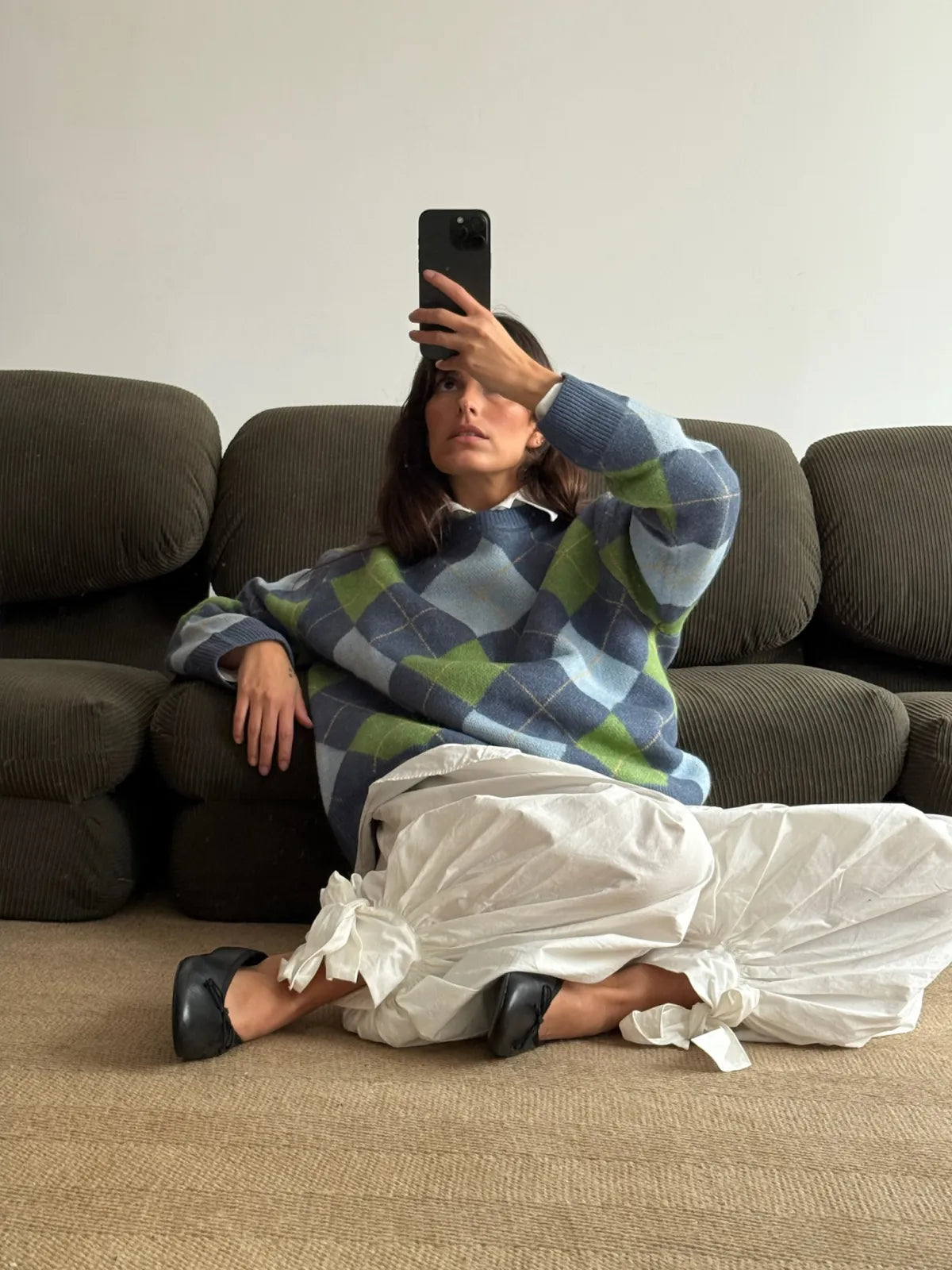 Person wearing blue and green checkered oversized sweater with white flowing pants and black shoes, sitting on tan couch