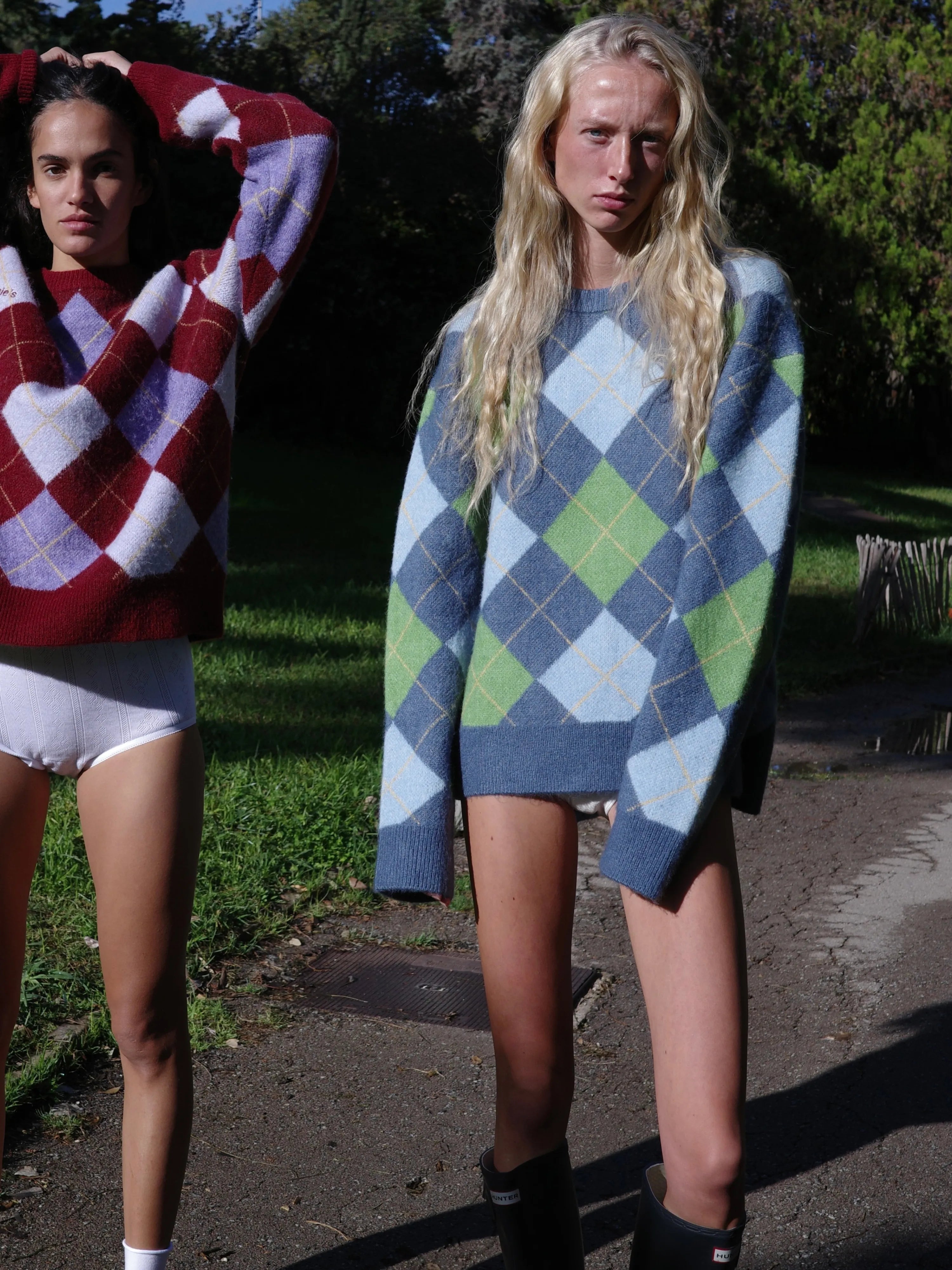 Two models wearing oversized argyle knit sweaters - one in red, purple and white, the other in blue, green and white diamonds.