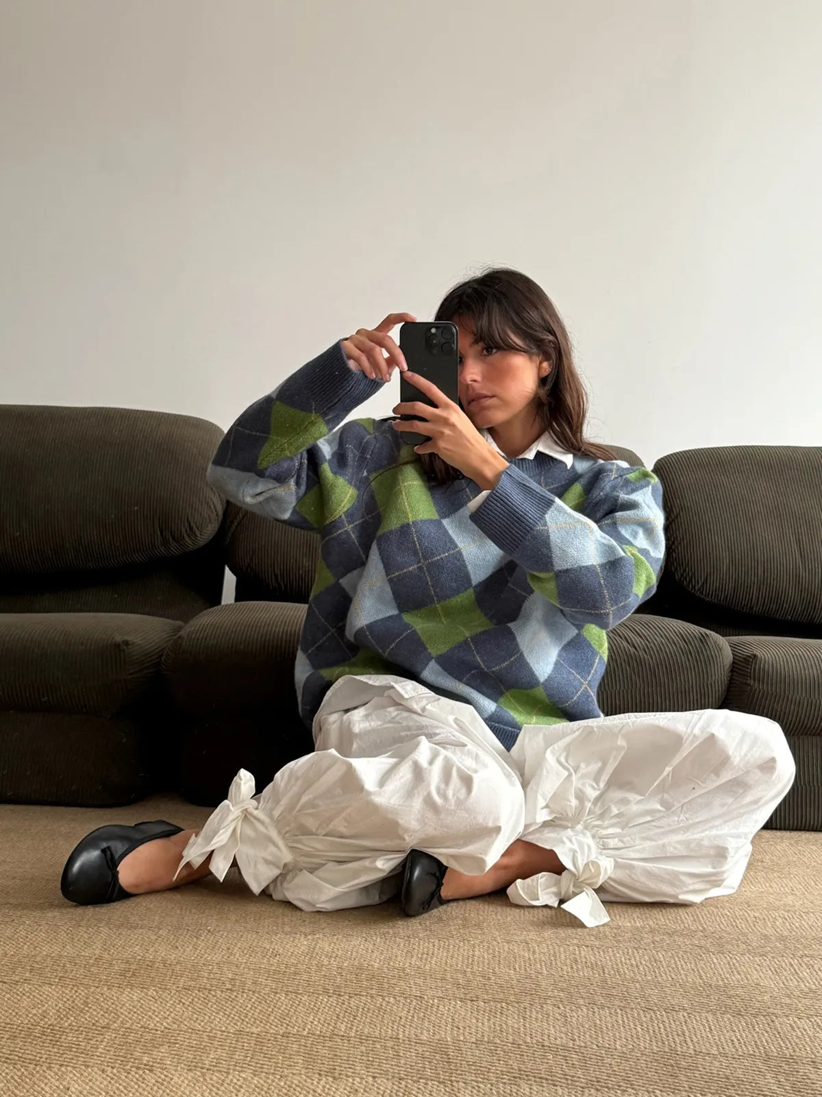Woman wearing oversized blue, green and gray argyle knit sweater with white pants, sitting on beige couch taking mirror selfie