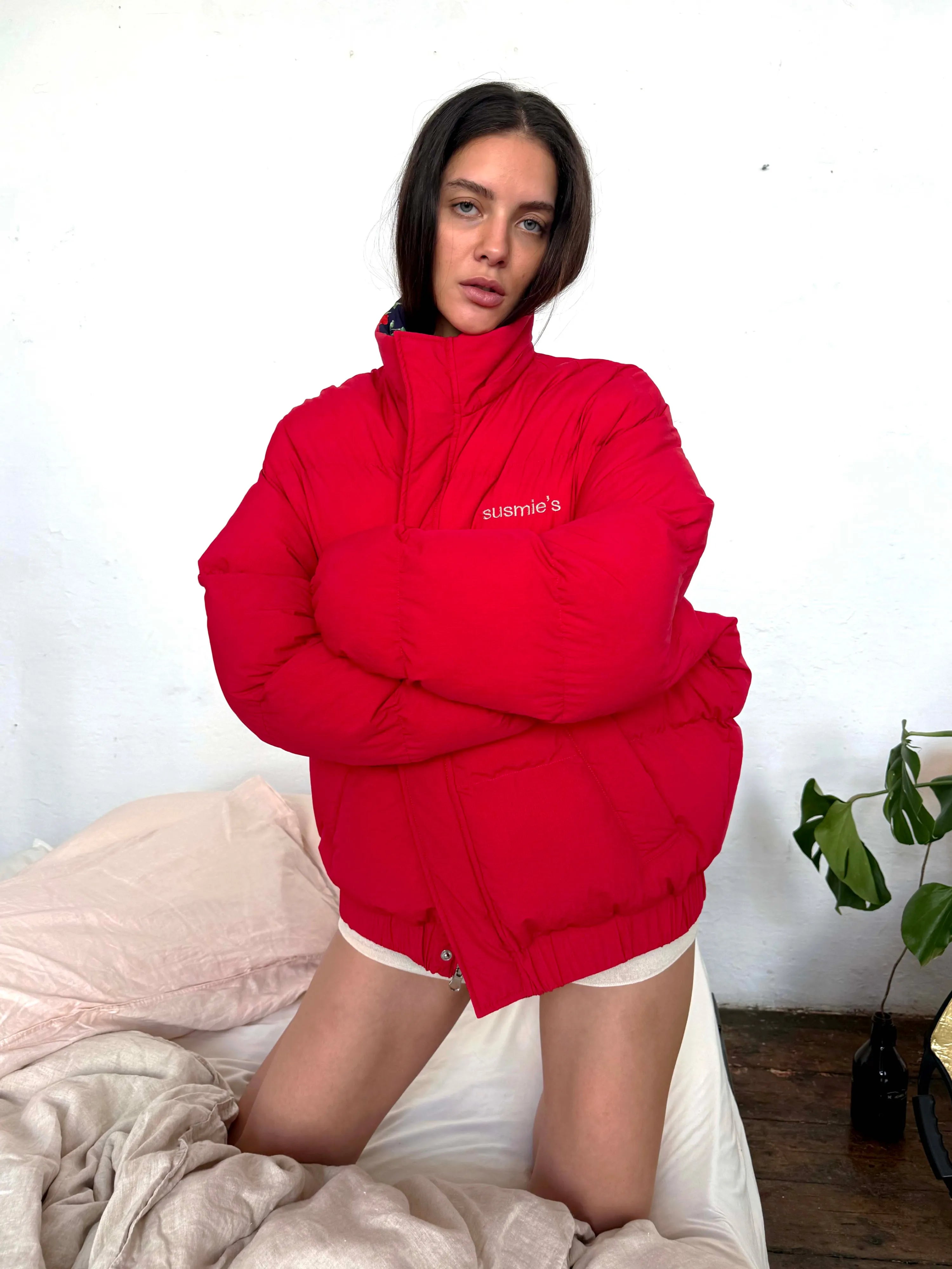 Red puffer jacket with susmie's embroidered logo, oversized fit, high collar, modeled on person kneeling on bed