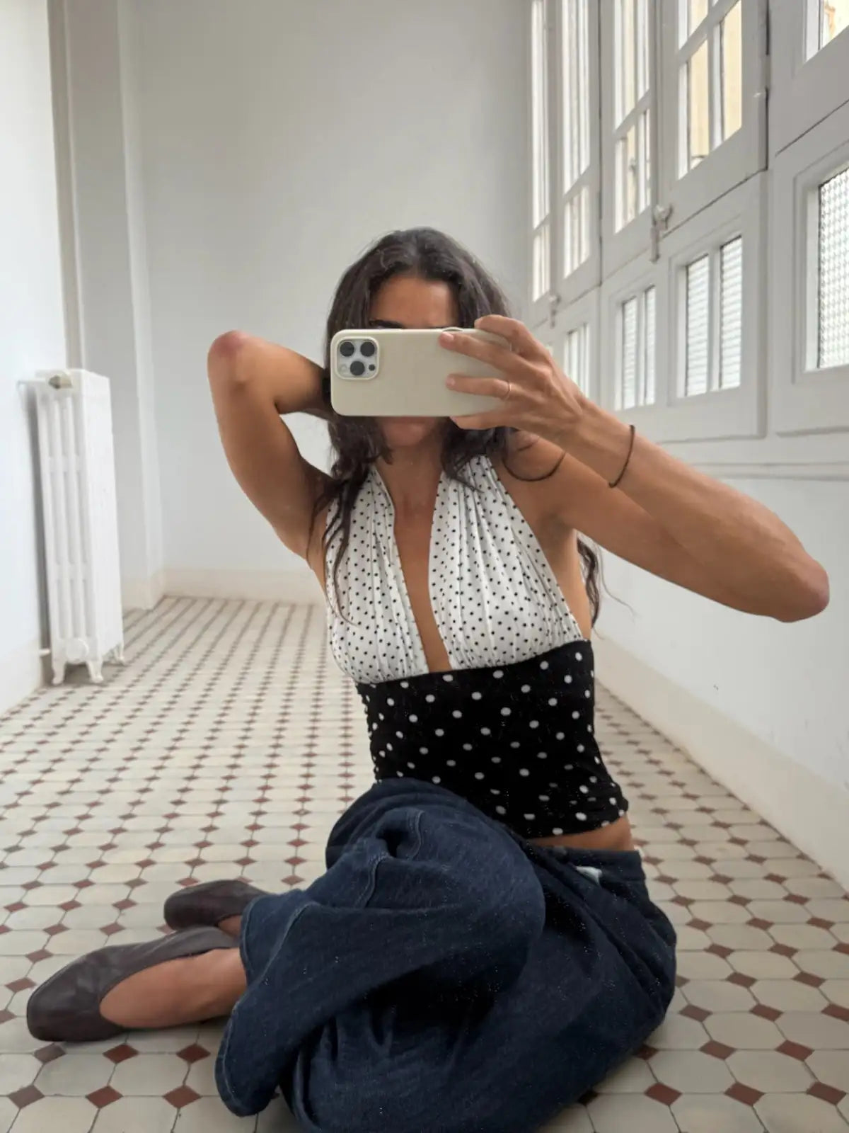 Woman wearing polka dot halter top and black bottoms sitting on vintage tile floor taking mirror selfie