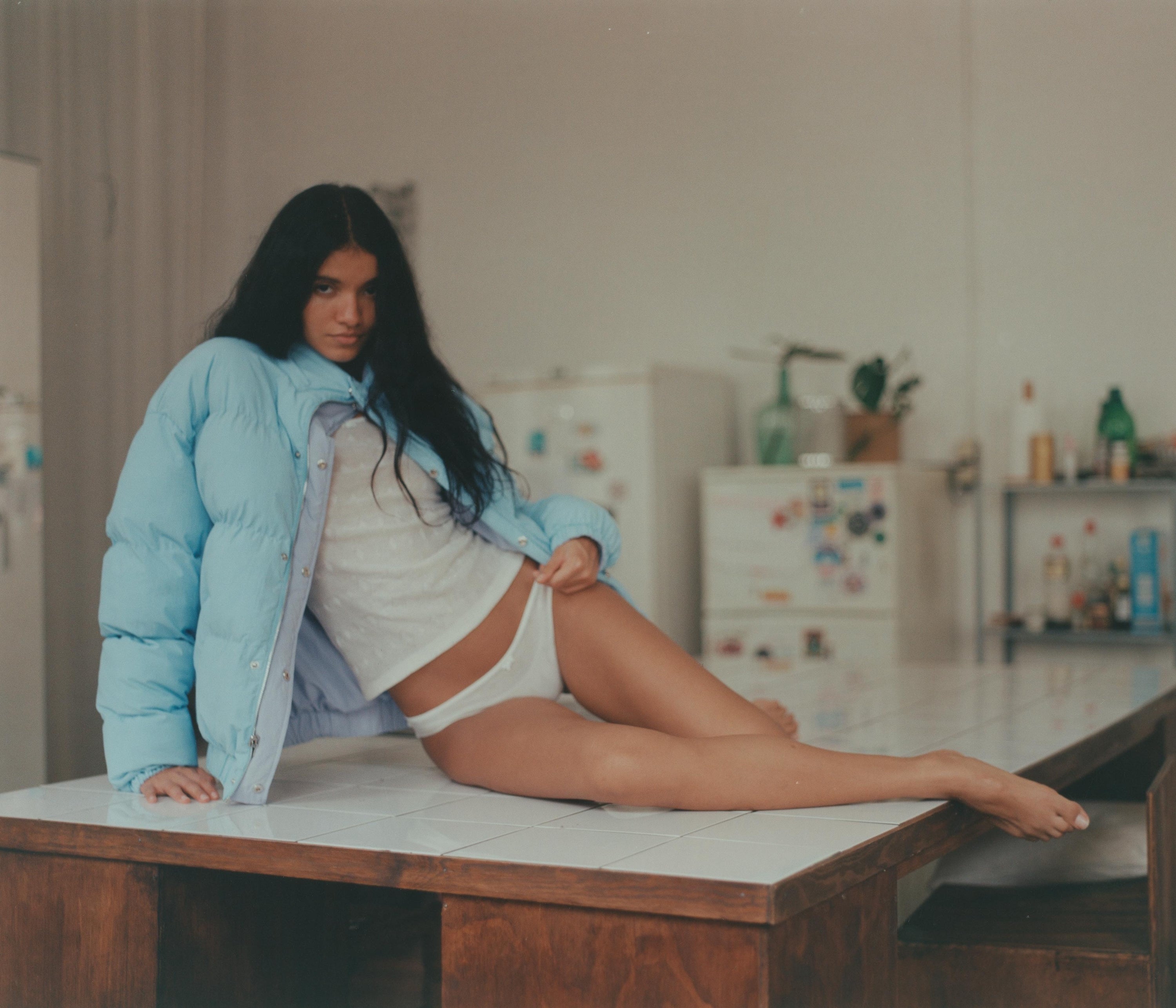 Woman in light blue puffer jacket and white crop top sitting on kitchen table in minimalist interior