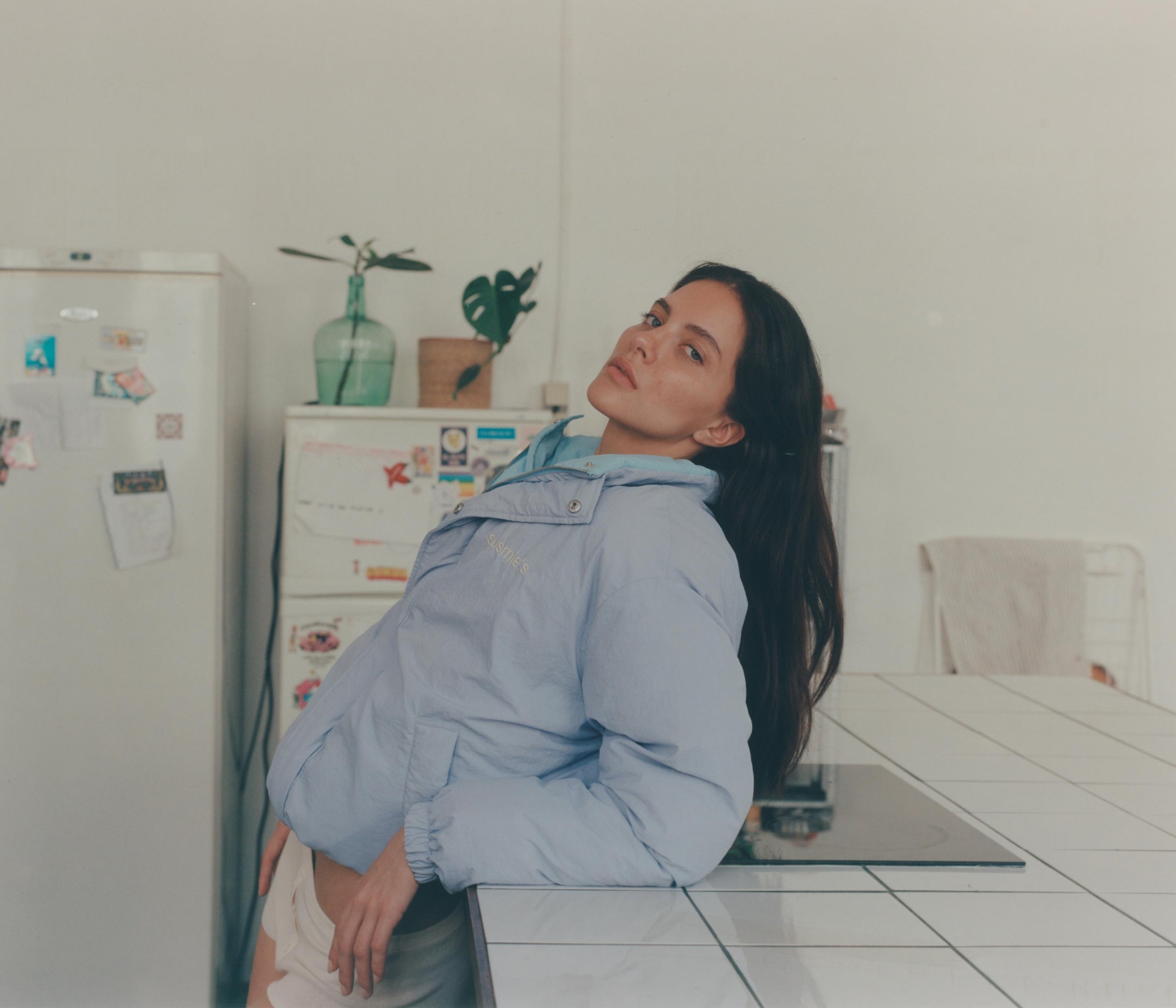 Woman wearing light blue oversized hooded jacket sitting on kitchen counter with long dark hair