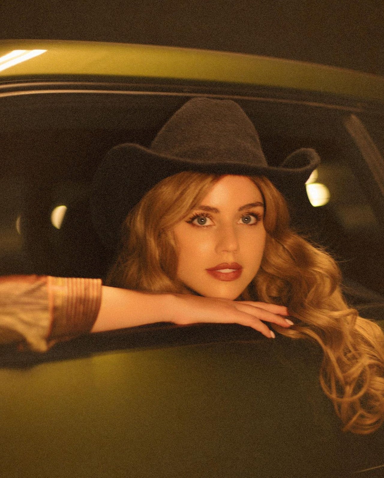 Woman with long wavy blonde hair wearing black hat leaning out of yellow taxi cab window at night