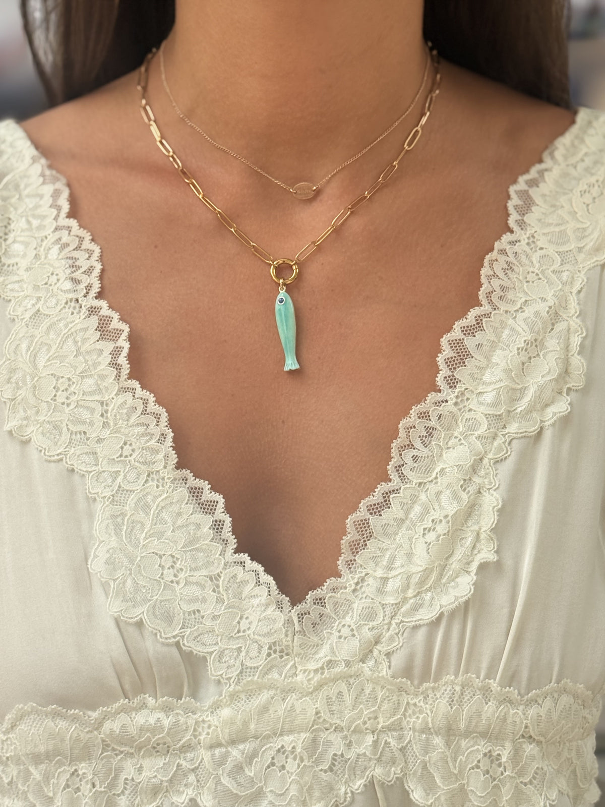 Gold layered necklace set with turquoise enamel fish charm pendant, worn over white lace top