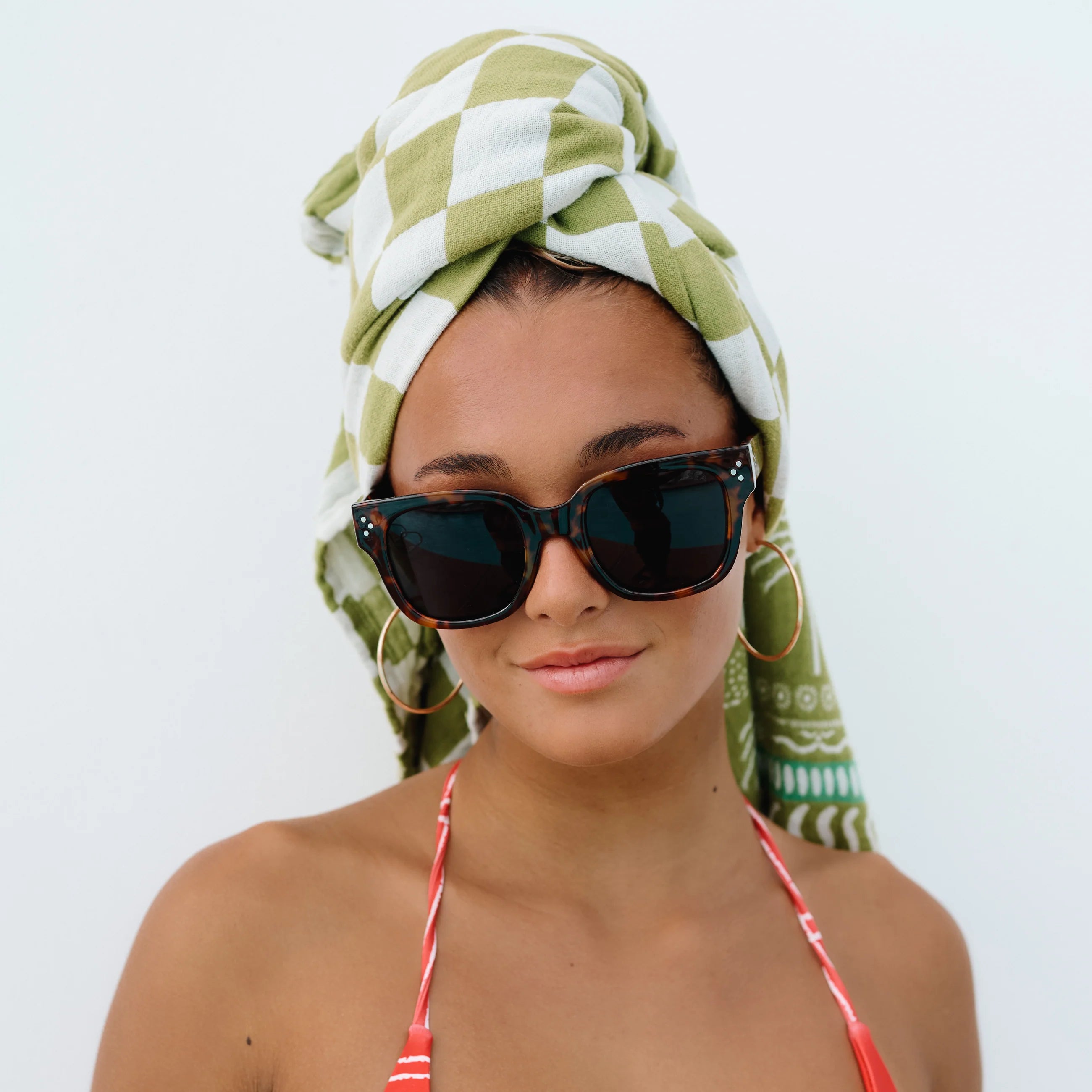 Woman wearing sunglasses and a green and white patterned headscarf on a plain background