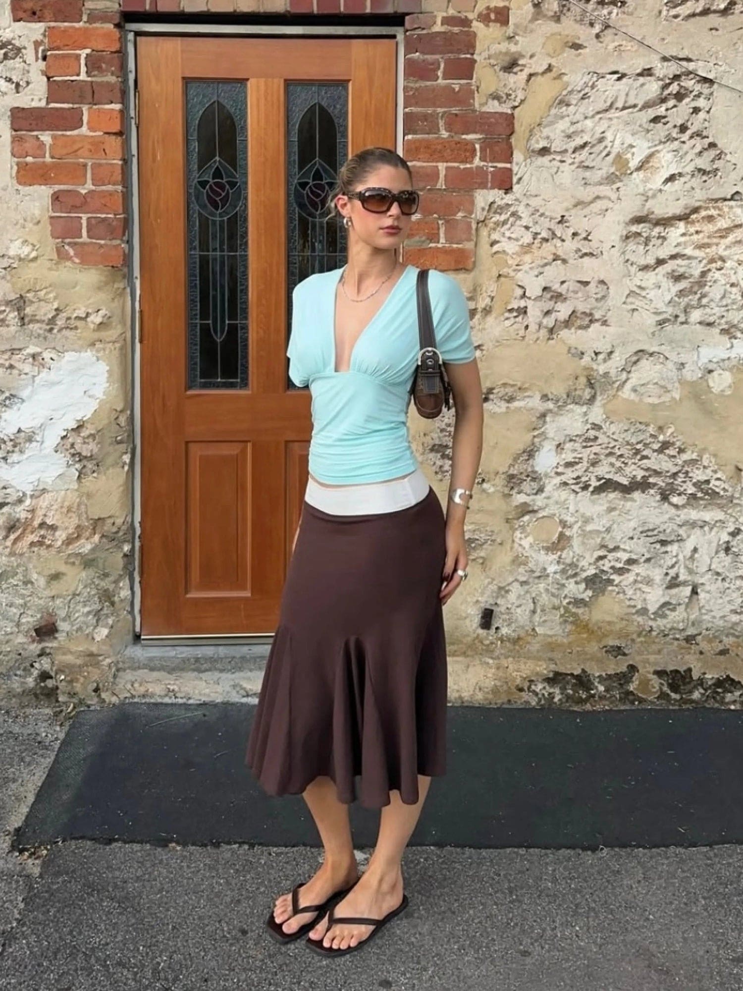 Woman wearing light blue wrap top, white underlayer, and brown midi skirt with sunglasses standing by weathered brick wall