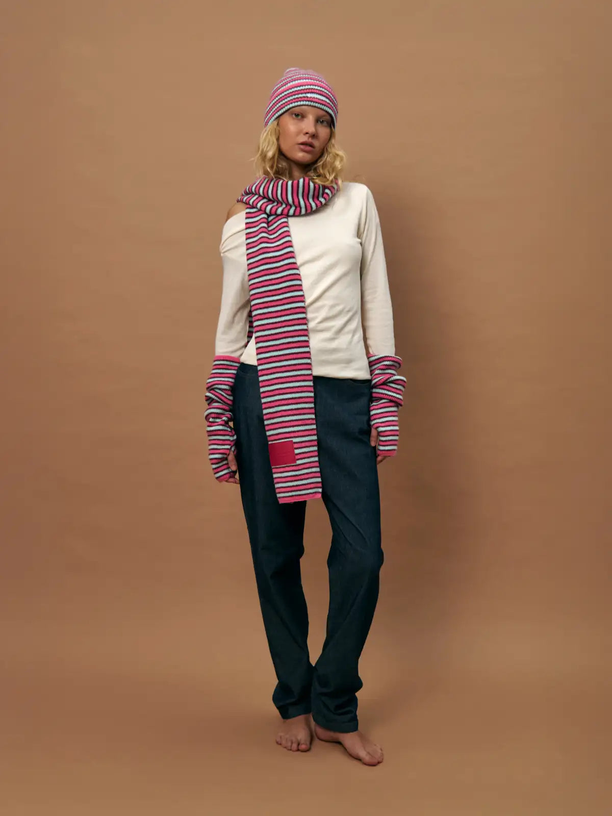 Woman wearing striped pink and navy knit beanie, matching scarf and arm warmers with cream sweater and dark pants