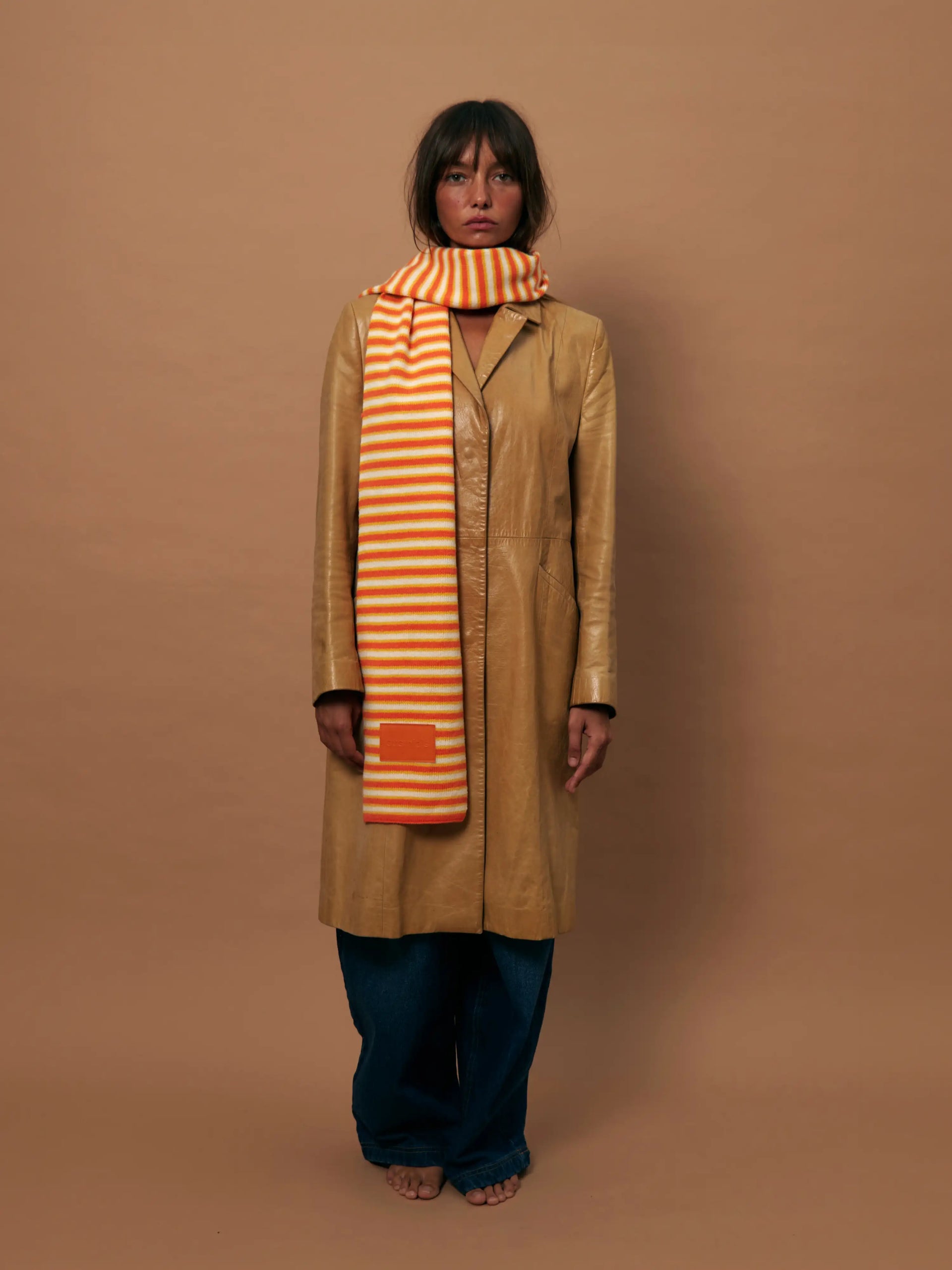 Tan leather coat with orange and cream striped scarf, worn with dark wide-leg jeans against beige background