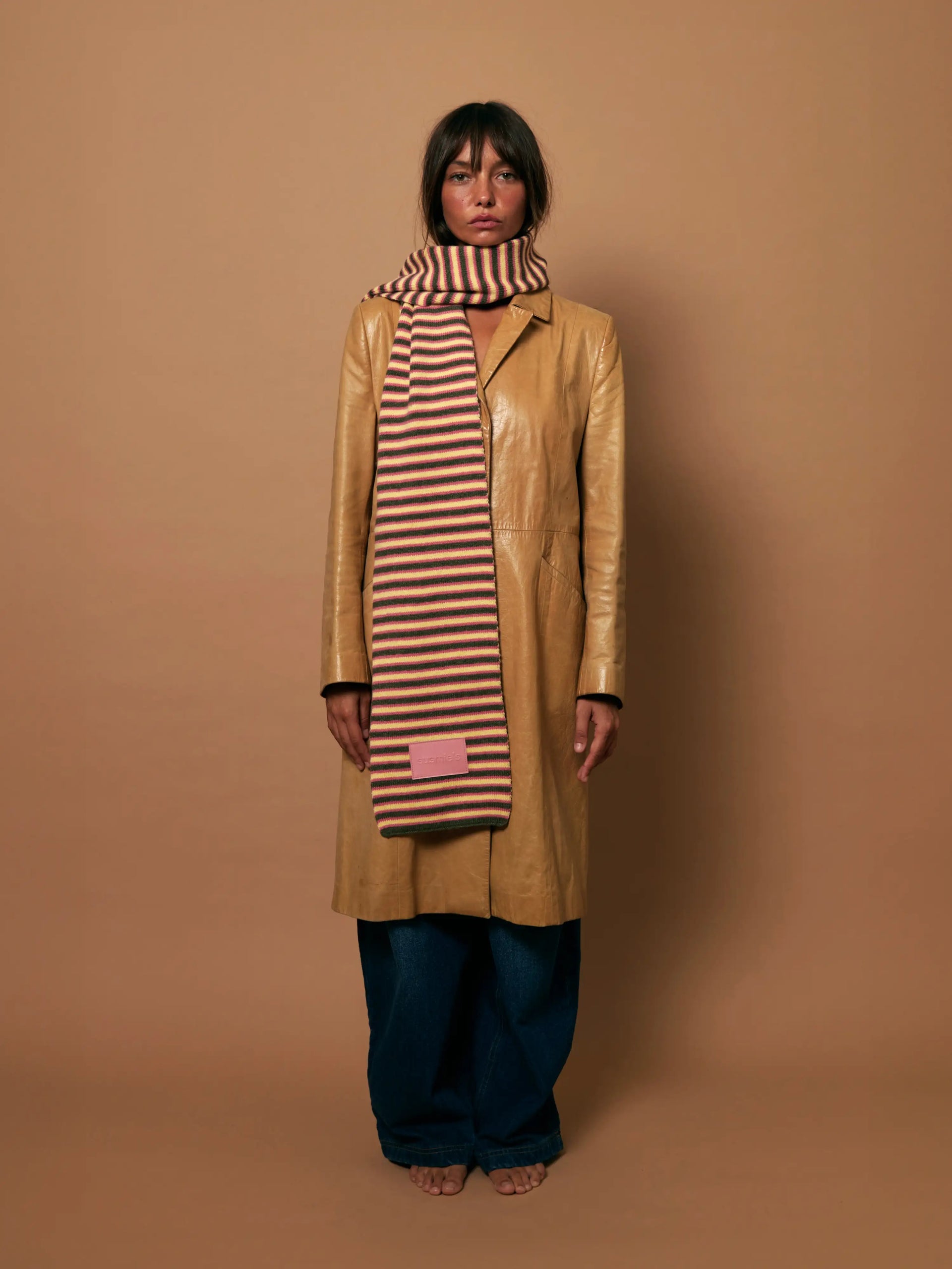 Tan leather coat with striped knit scarf in beige and brown, worn over wide-leg blue jeans against neutral background