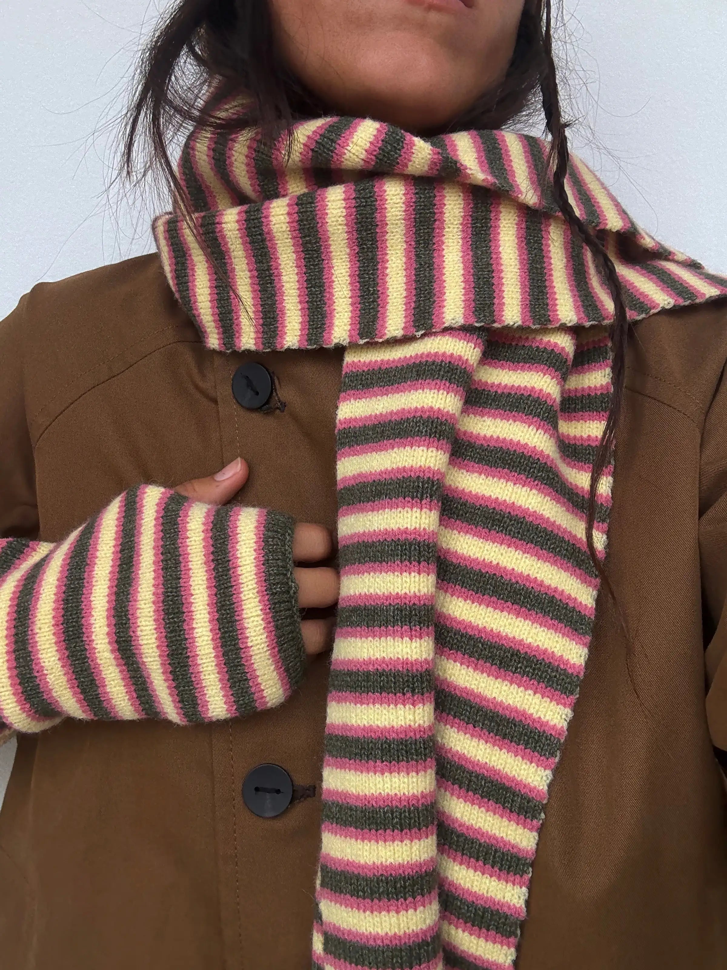 Striped knit scarf in pink, yellow, and charcoal gray worn with matching fingerless gloves and brown coat