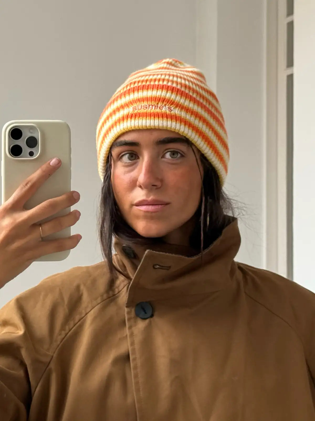 Orange and cream striped knit beanie hat with embroidered text, worn by person in brown jacket taking mirror selfie