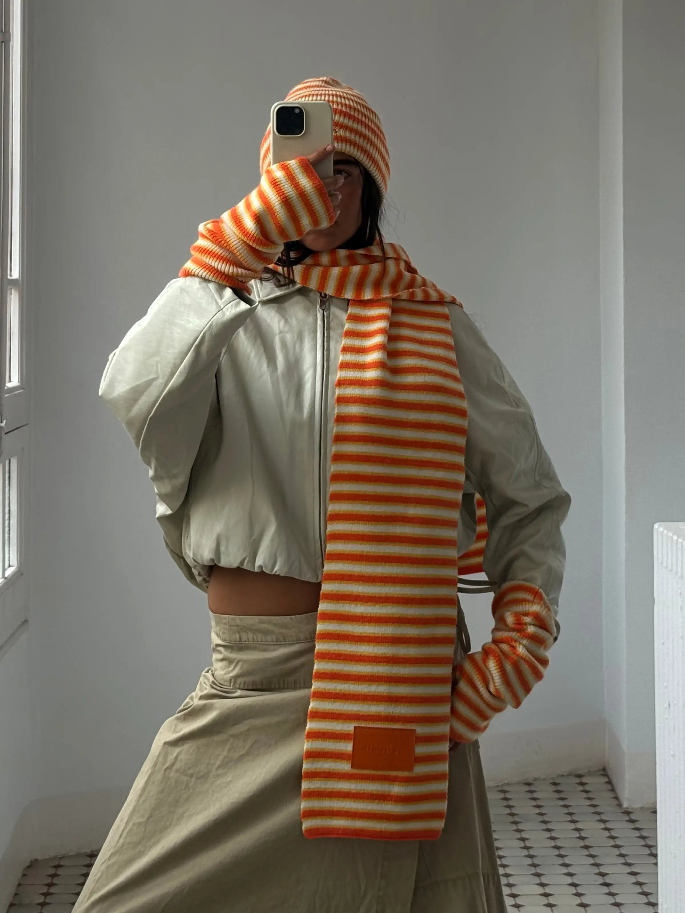 Orange and cream striped knit scarf and beanie set worn with beige jacket, mirror selfie showing coordinated winter accessories