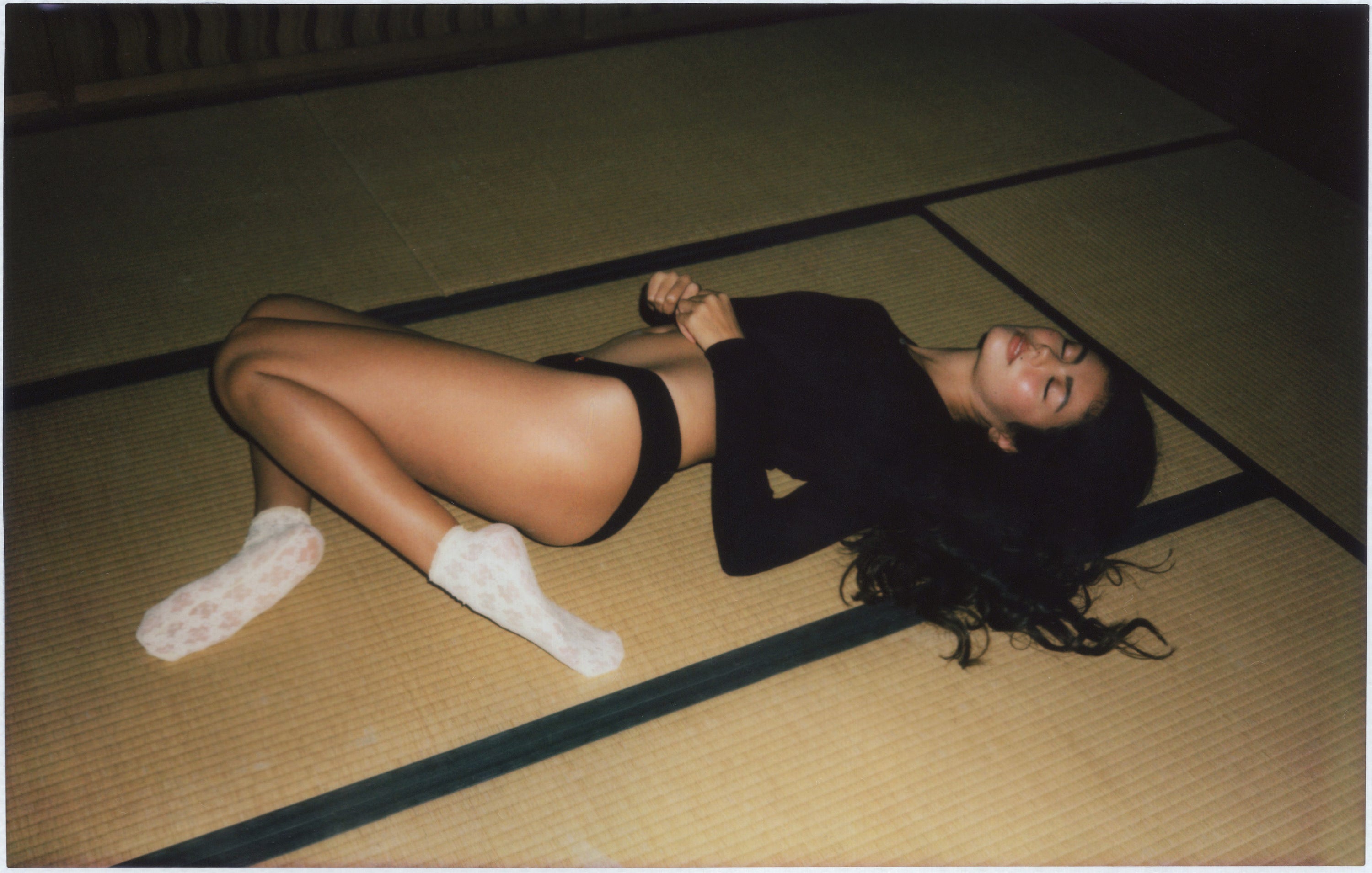 Woman lying on tatami mat wearing black bodysuit and white fuzzy slippers in Japanese-style room