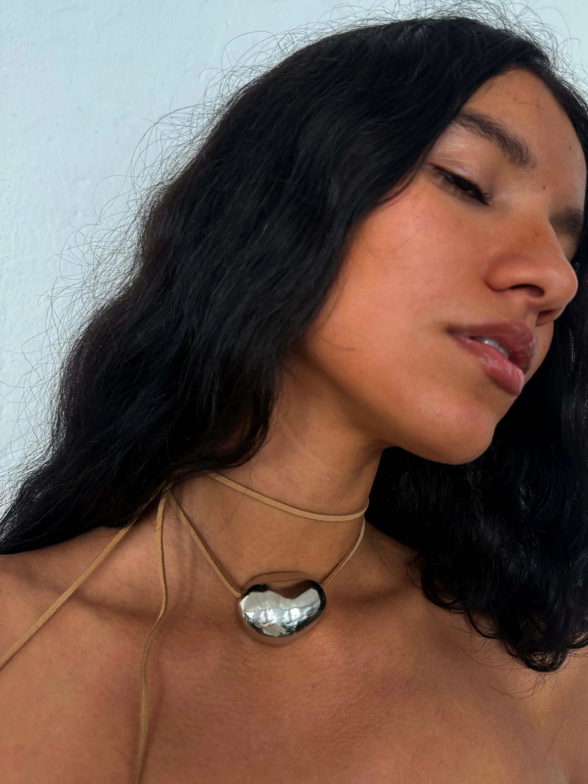 Silver heart-shaped pendant necklace on tan leather cord worn by woman with black hair against blue background