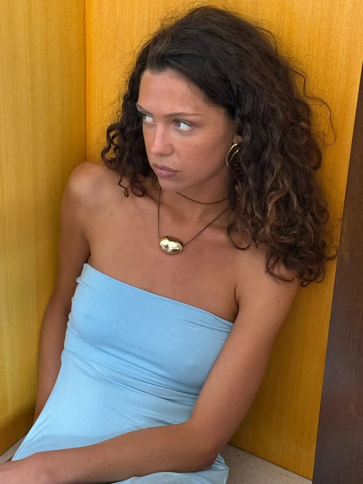 Woman wearing light blue strapless tube dress with gold pendant necklace and hoop earrings against yellow wall