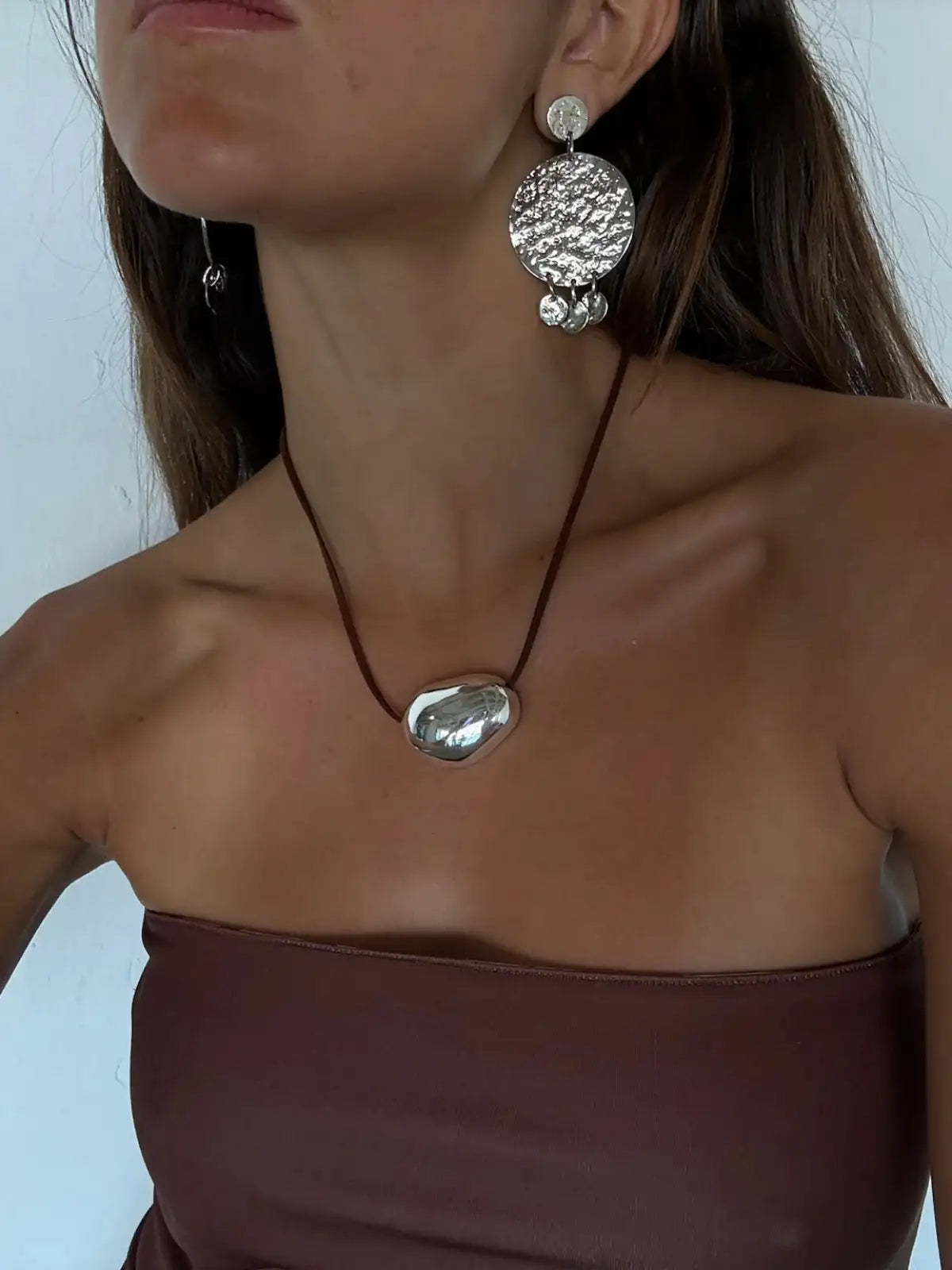 Silver hammered disc drop earrings and matching oval pendant necklace on brown cord against brown strapless top