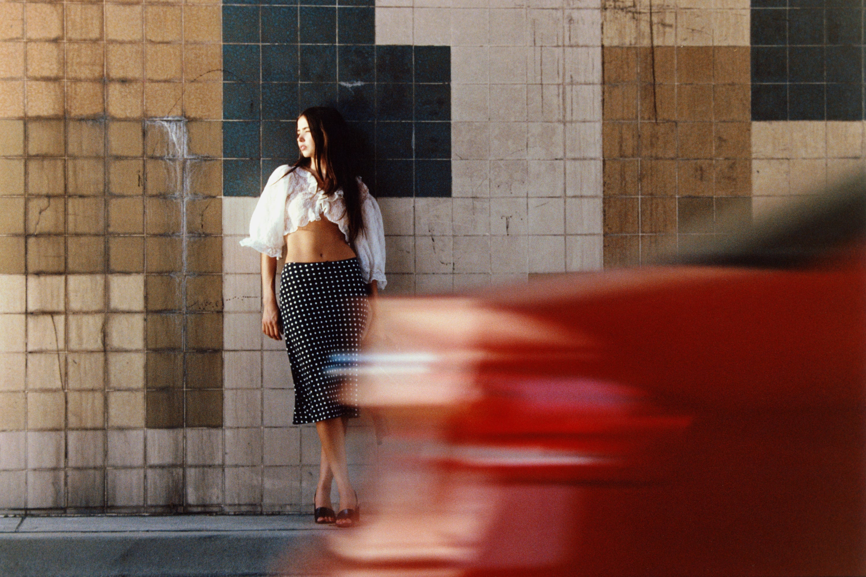 Woman in white cropped lace top and black polka dot midi skirt standing against colorful tiled wall with motion blur