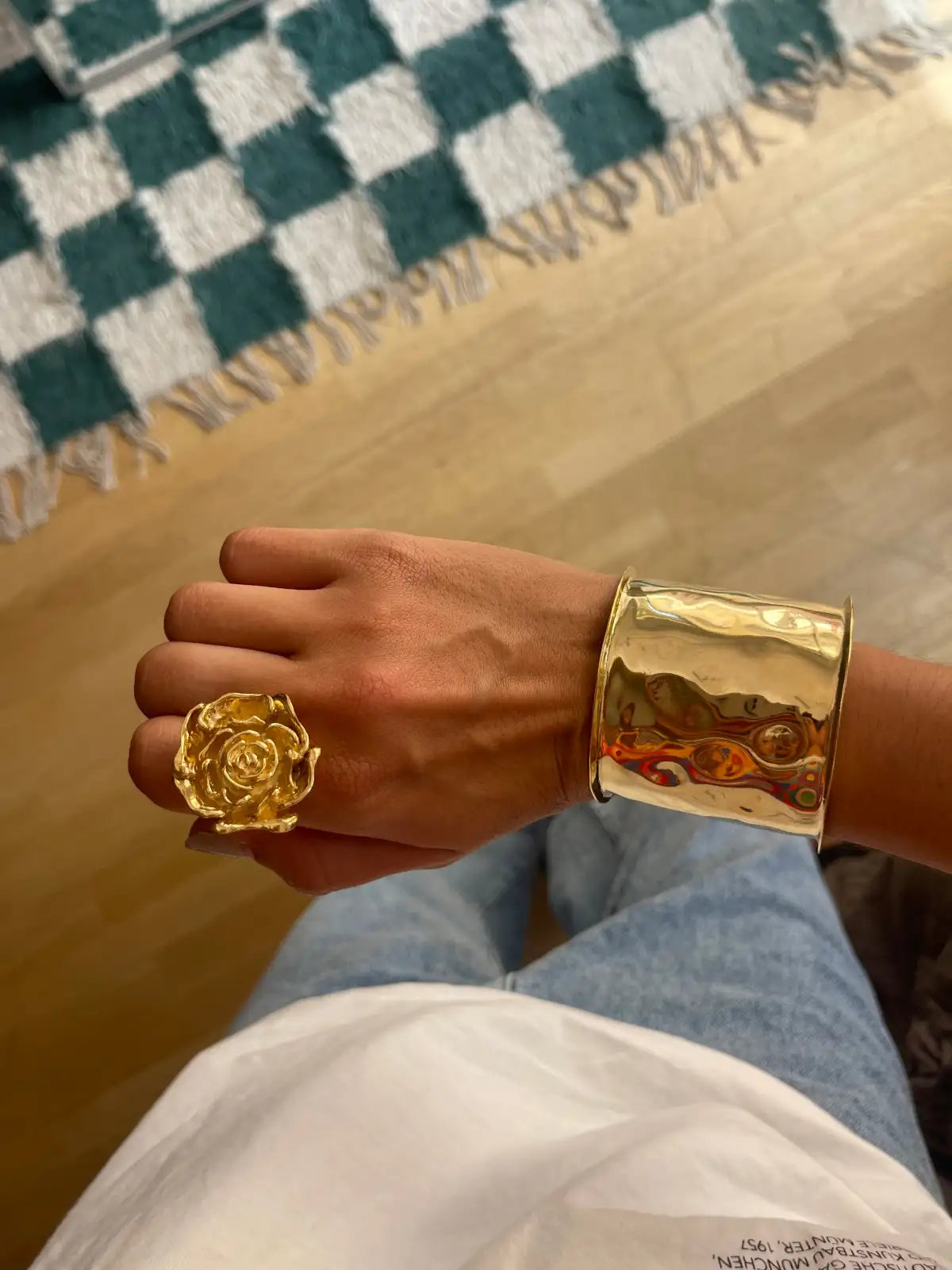 Gold hammered metal cuff bracelet and rose ring set on hand, showing textured metallic finish against light wood floor