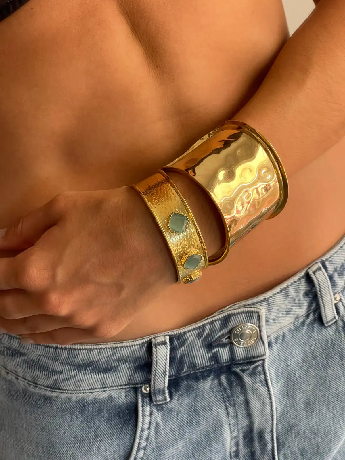 Gold metallic cuff bracelets stacked on wrist, featuring wide polished band and textured band with blue stone accents