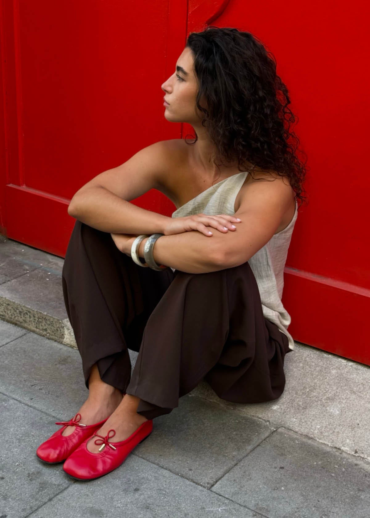Red leather ballet flats with bow detail and gold hardware, shown on model seated against red backdrop.