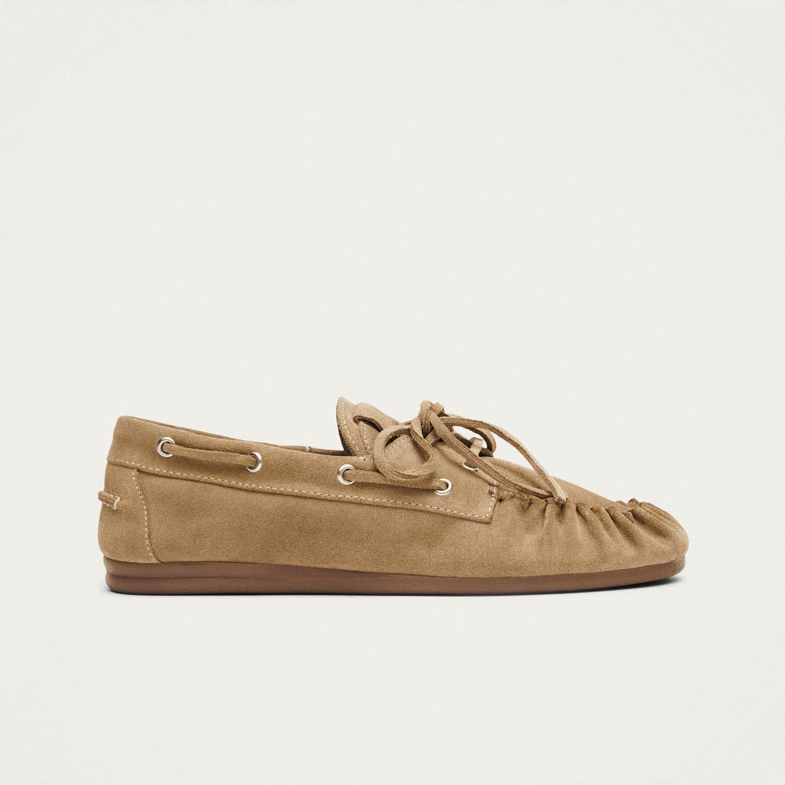 Tan suede moccasin loafer with leather laces and brown rubber sole, side view on beige background