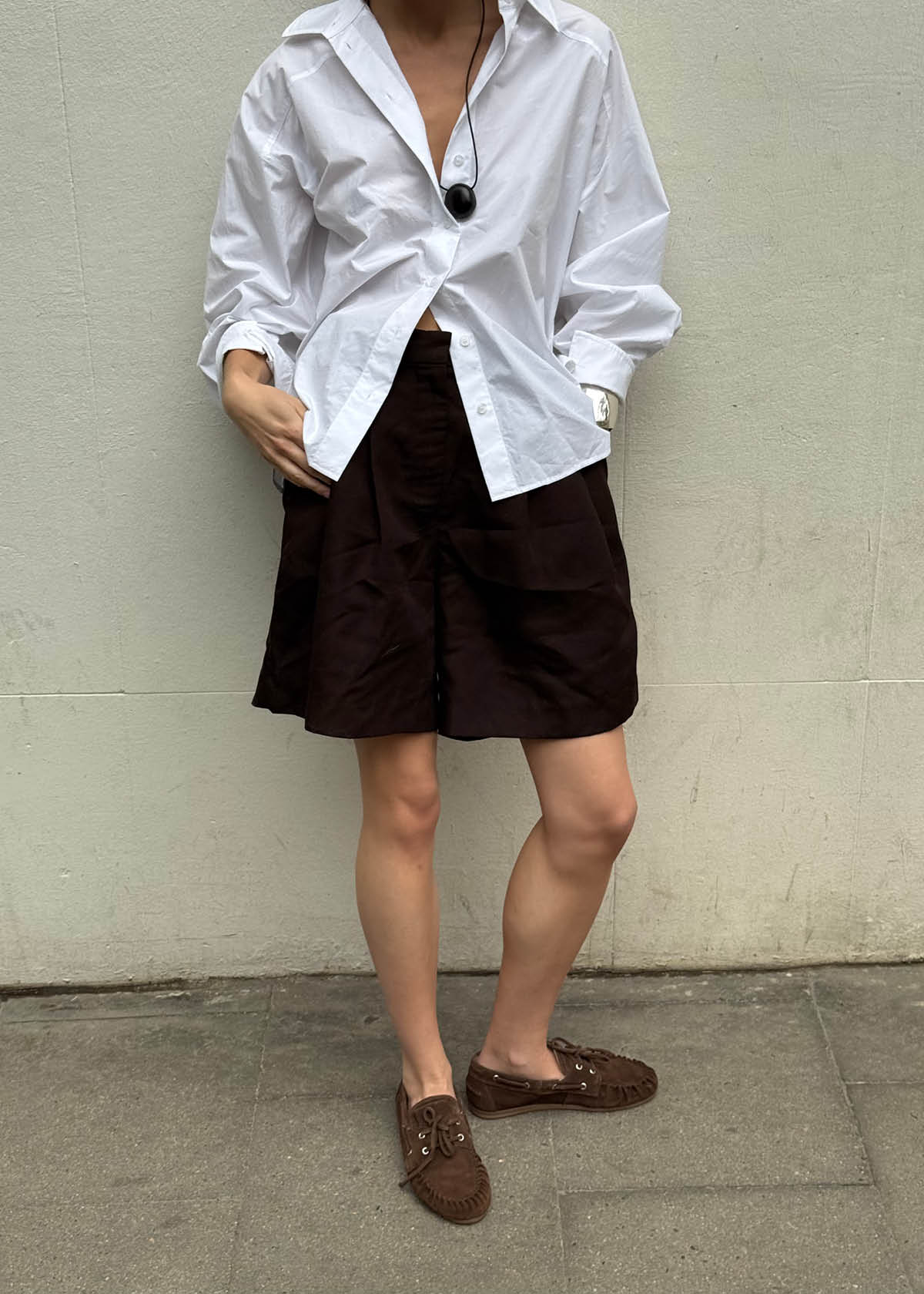Person wearing oversized white button-up shirt with brown shorts and matching brown suede loafers against neutral wall