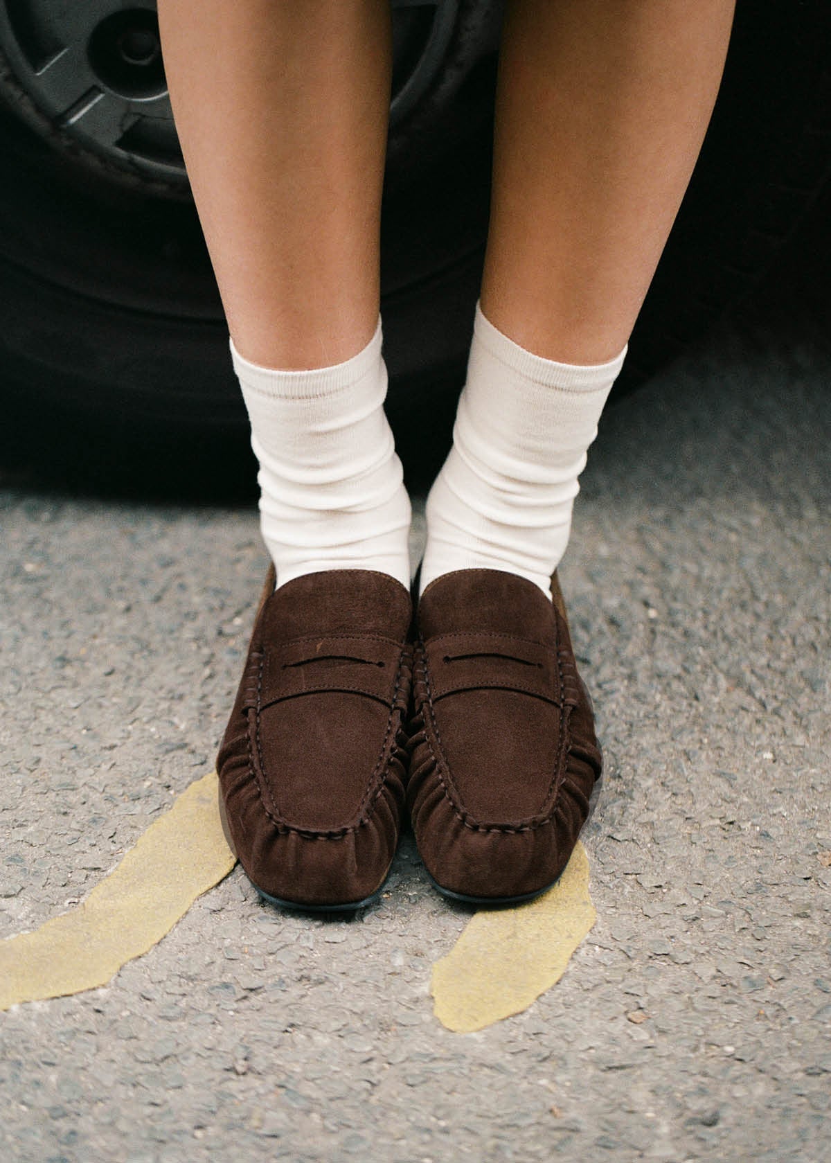 Brown suede moccasin loafers with white ribbed socks, shown on legs standing on pavement