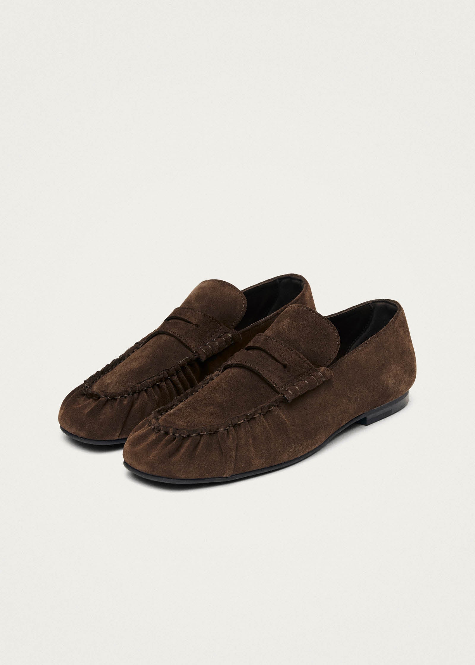 Brown suede penny loafers with moc-toe stitching and black leather lining on beige background