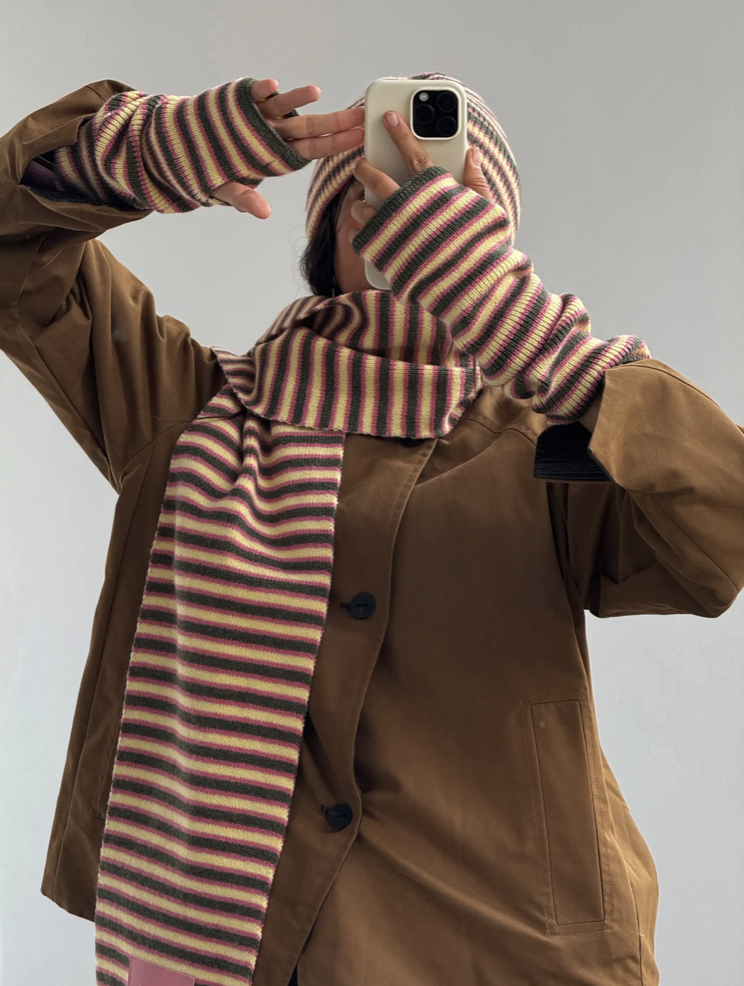Person wearing brown coat with multicolored striped knit scarf and matching fingerless gloves taking mirror selfie