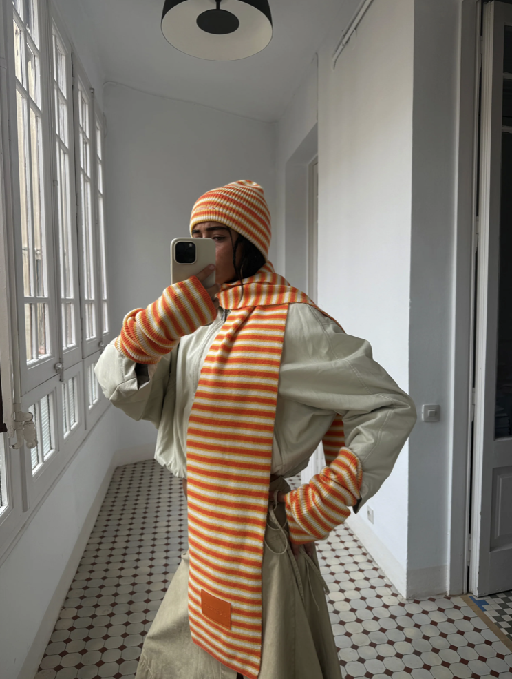 Person wearing orange and white striped knit beanie and matching scarf with cream coat in bright hallway
