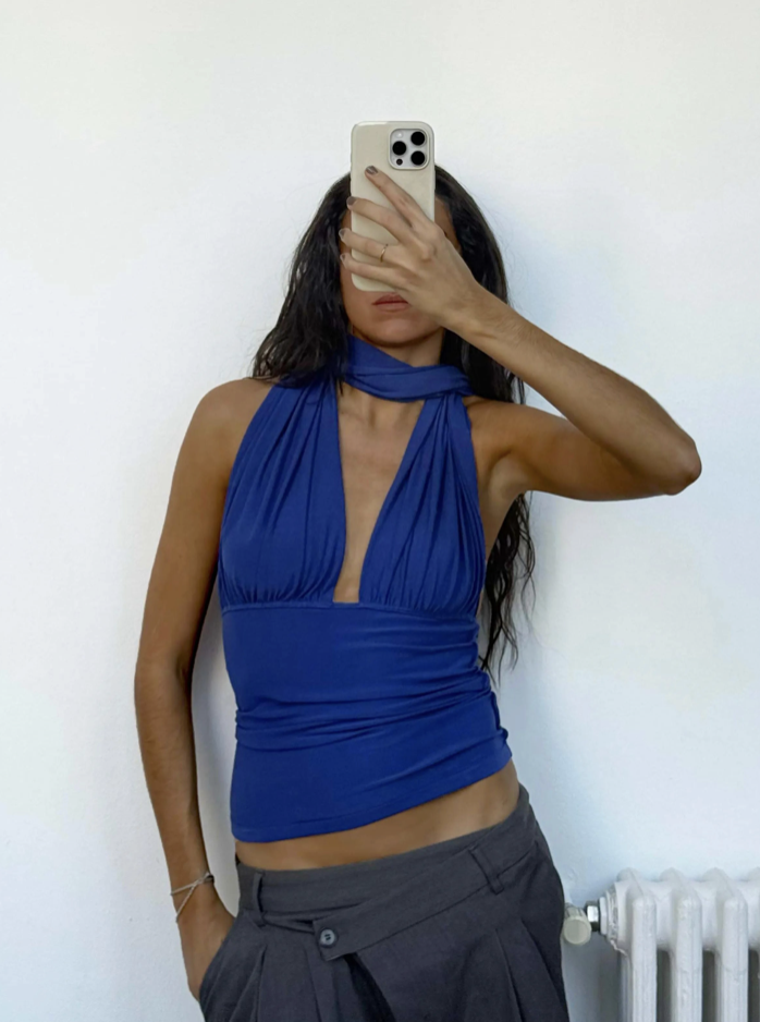 Blue halter neck crop top with deep V-neckline and ruched waist detail, worn with gray low-rise pants