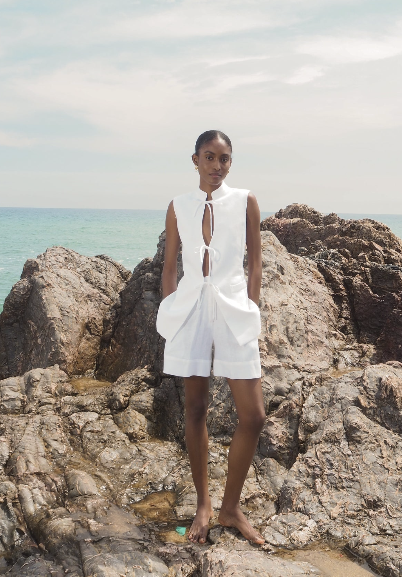 White sleeveless linen top with collar and tie detail, paired with matching white shorts, modeled on rocky coastline