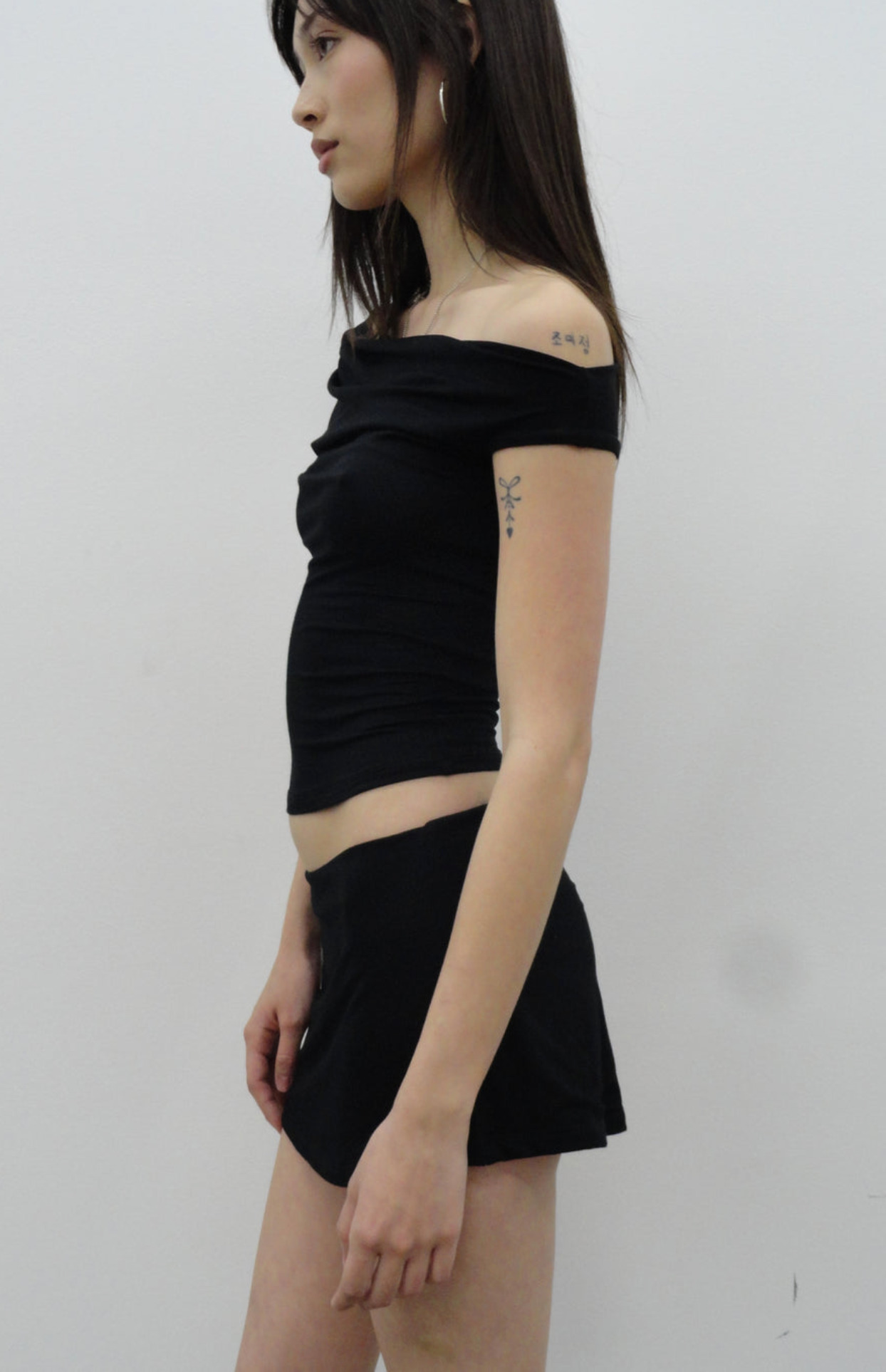 Woman wearing black off-shoulder crop top and black mini skirt, showing side profile with visible tattoos on shoulder and arm