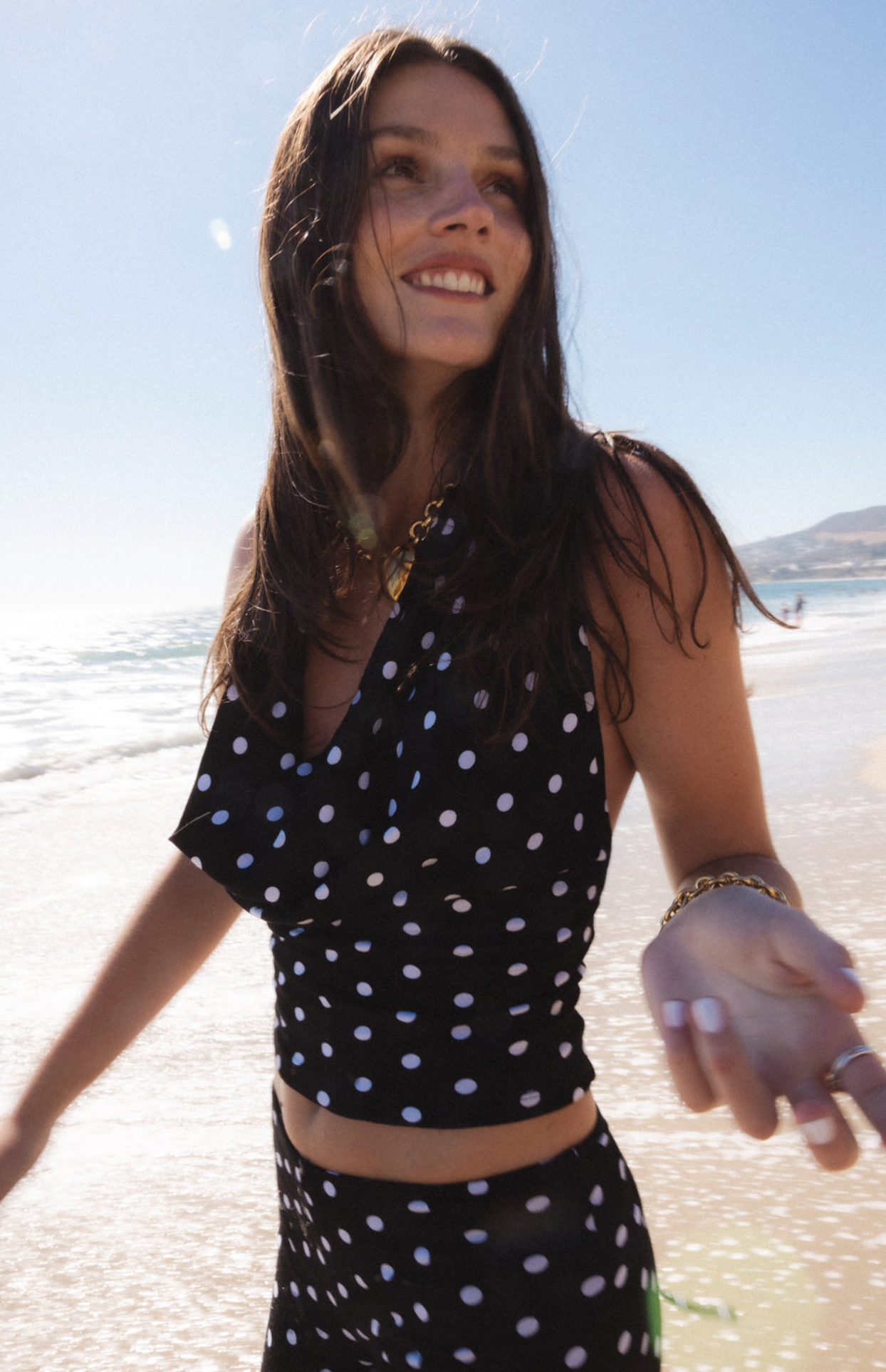 Woman on beach wearing black polka dot crop top and skirt set with gold chain necklace and bracelet