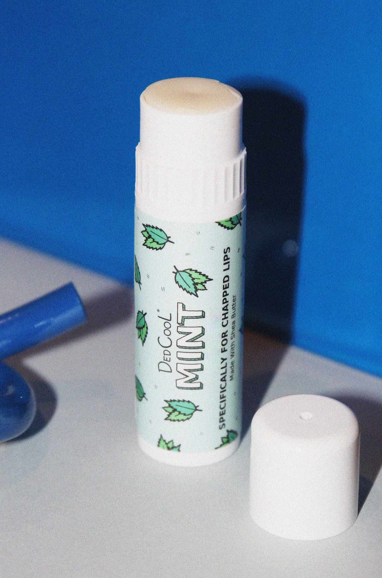 White lip balm tube with mint green label featuring leaf pattern, cap removed and placed beside it on white surface