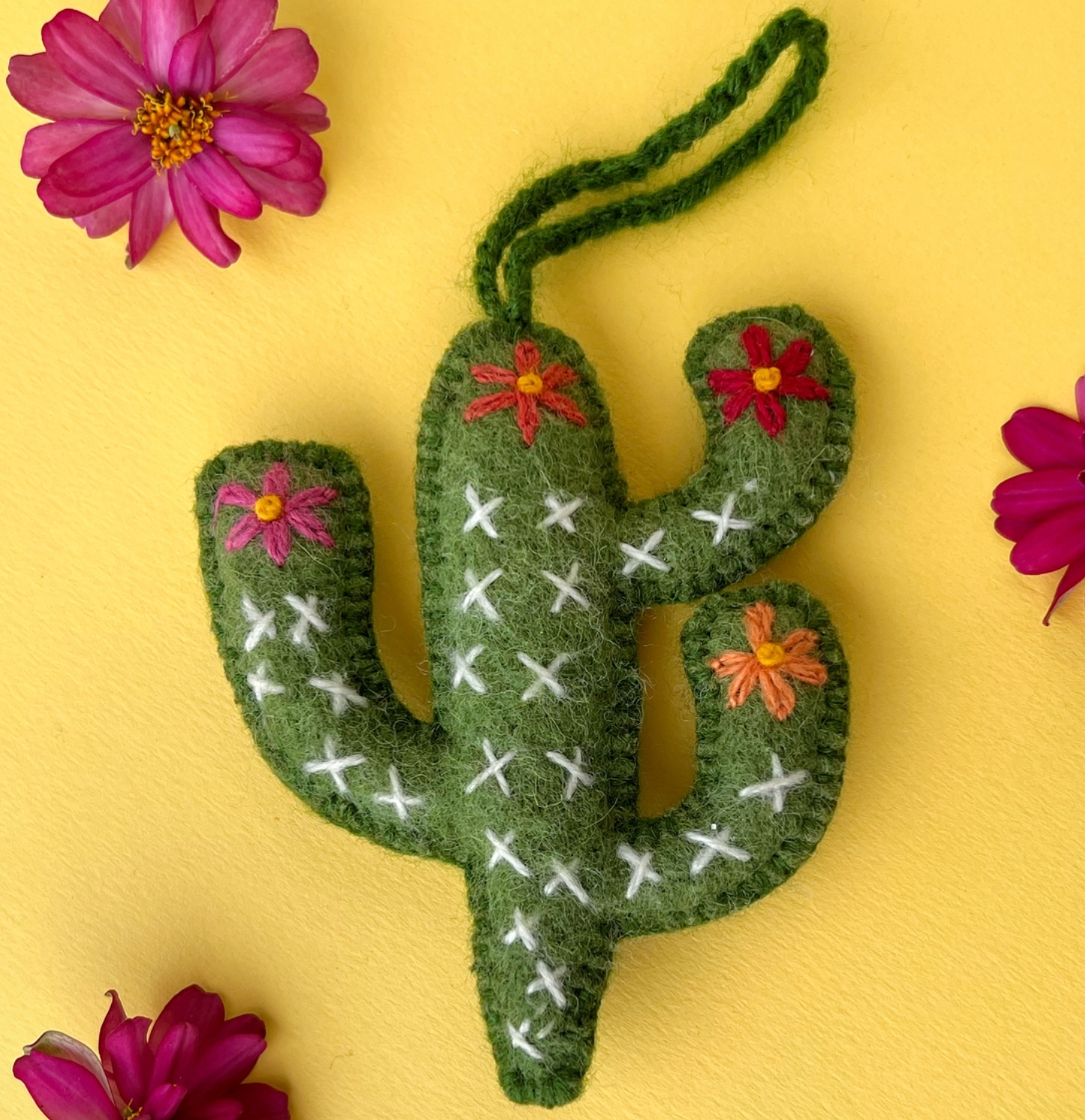 Green felt cactus ornament with white X stitching and colorful embroidered flowers on yellow background with pink blooms