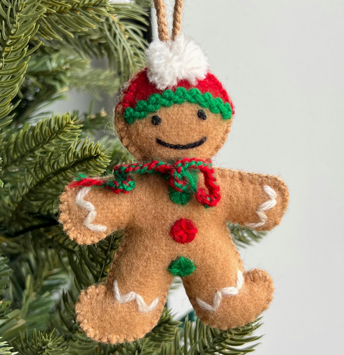 Felt gingerbread man ornament with red and green scarf, white pom-pom hat, and embroidered smile hanging on Christmas tree