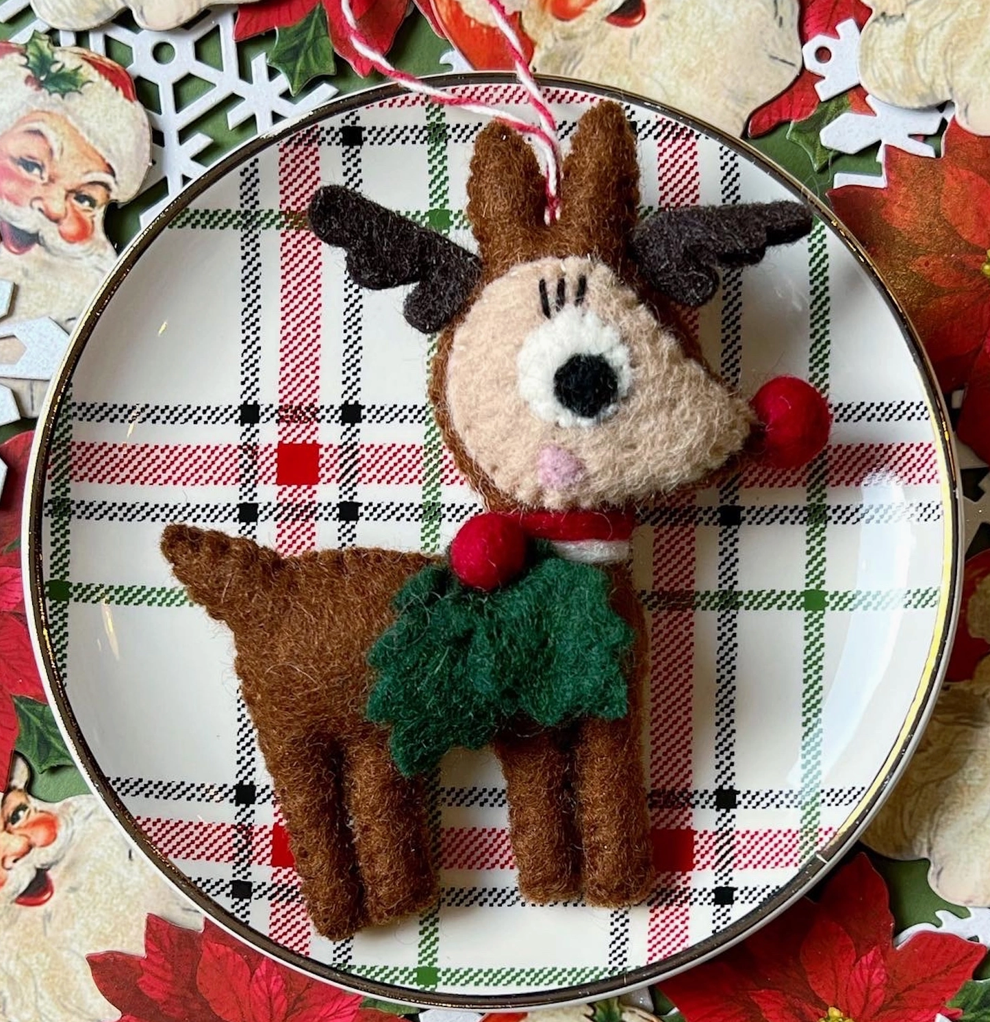 Felt reindeer ornament in brown with black antlers, green sweater, and candy cane string on plaid holiday plate